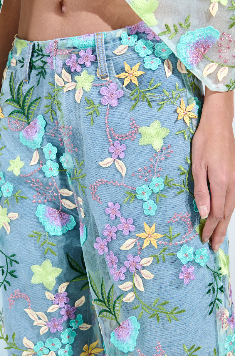A close-up showcases a person in high-rise Gardenia Denim with Floral Tulle Wide Leg Jeans, adorned with vibrant pink, yellow, green, and blue floral embroidery. The design extends to the left sleeve, beautifully accentuating the jeans' light blue background. 5