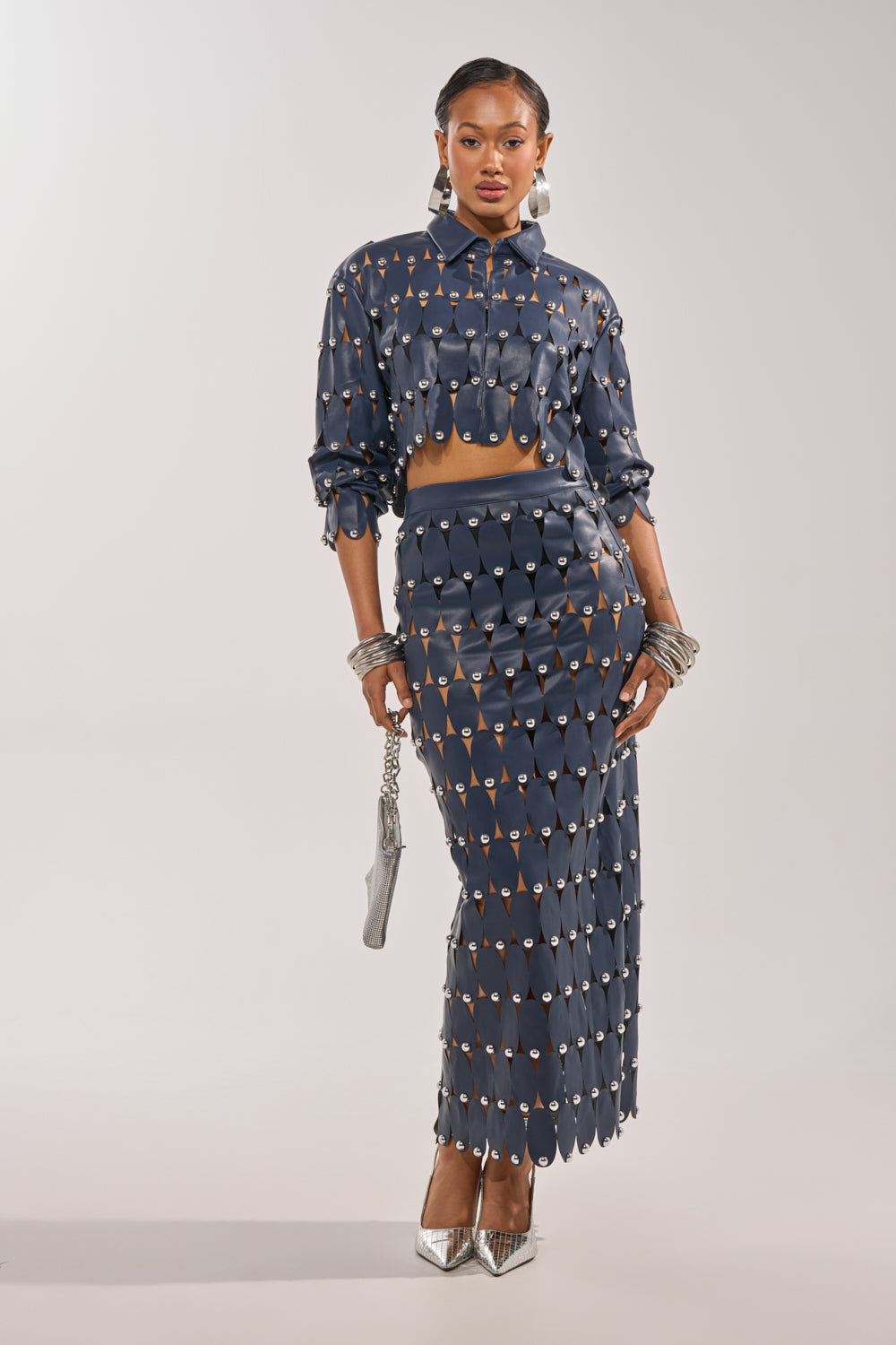 A woman poses in THE SNEAK PEEK STUDDED PU MIDI SKIRT IN NAVY, featuring teardrop cutouts and silver hardware. She pairs it with matching silver heels, a small silver bag, bold silver jewelry, and her hair slicked back. 0