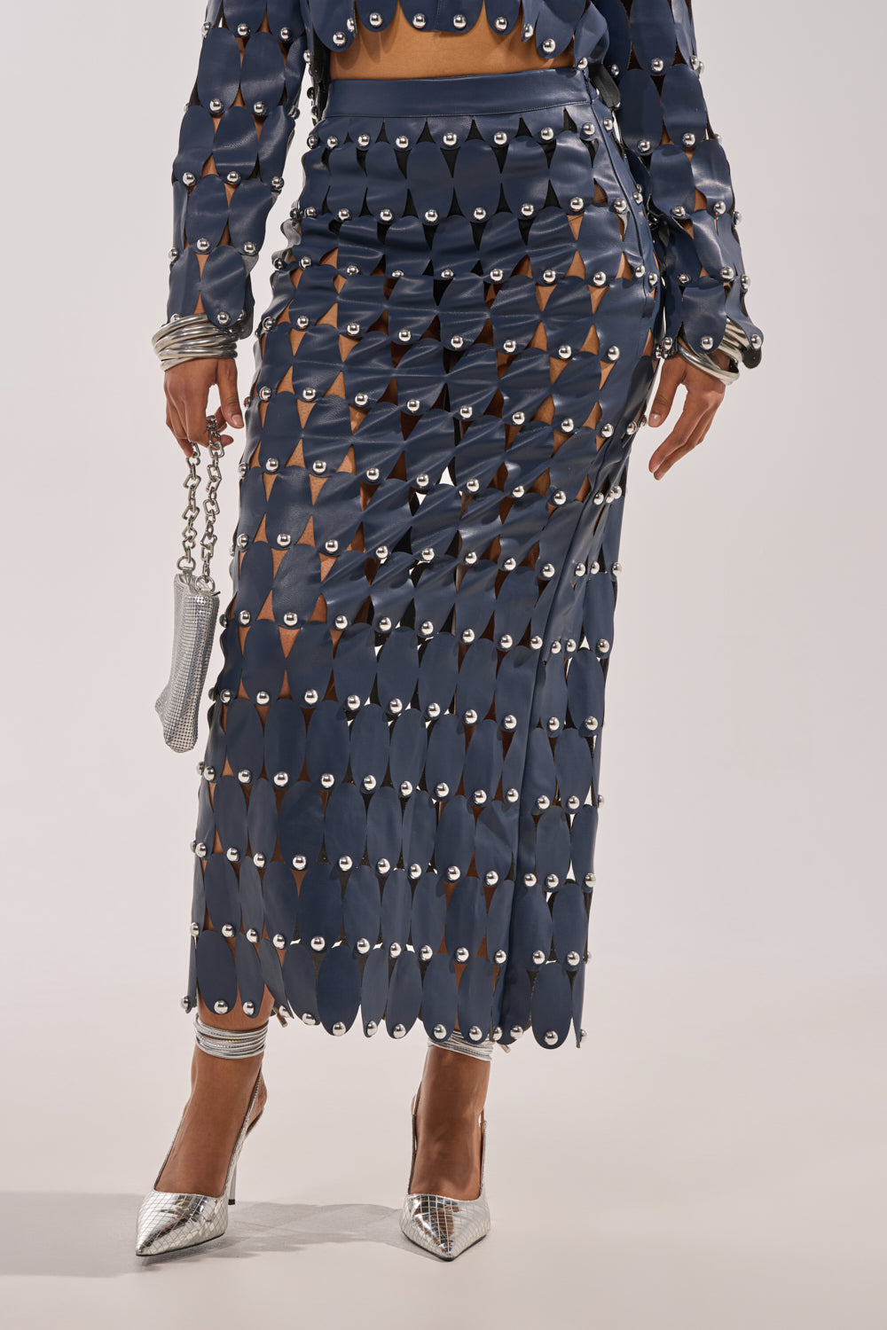 Wearing THE SNEAK PEEK STUDDED PU MIDI SKIRT IN NAVY with geometric cutouts and silver accents, paired with a matching top, metallic purse, and pointed silver heels; the person’s face is not visible. 2
