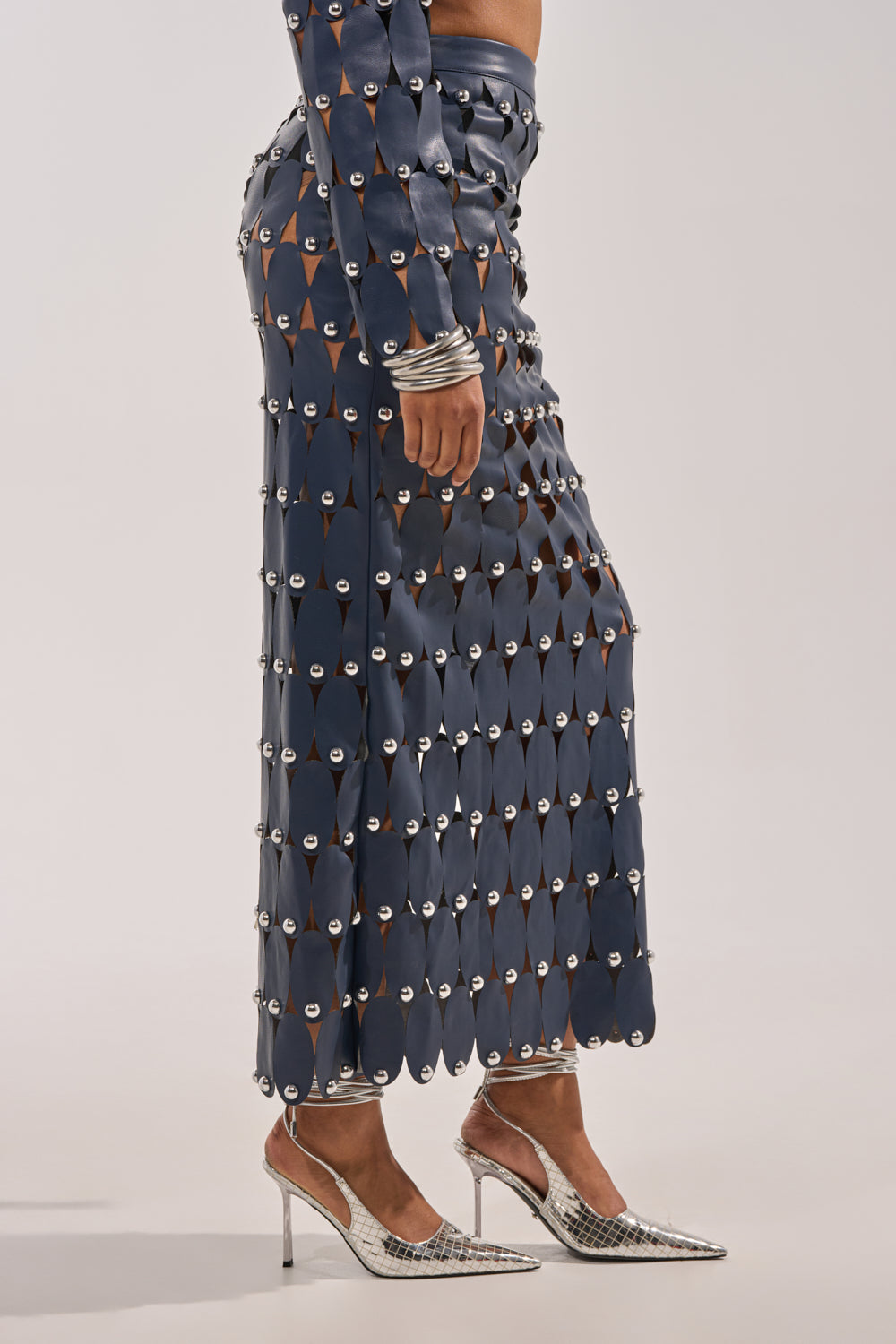 A person is shown from the shoulders down, wearing THE SNEAK PEEK STUDDED PU MIDI SKIRT IN NAVY with circular metallic cutouts, silver bracelets, and silver pointed high heels against a plain background. 3