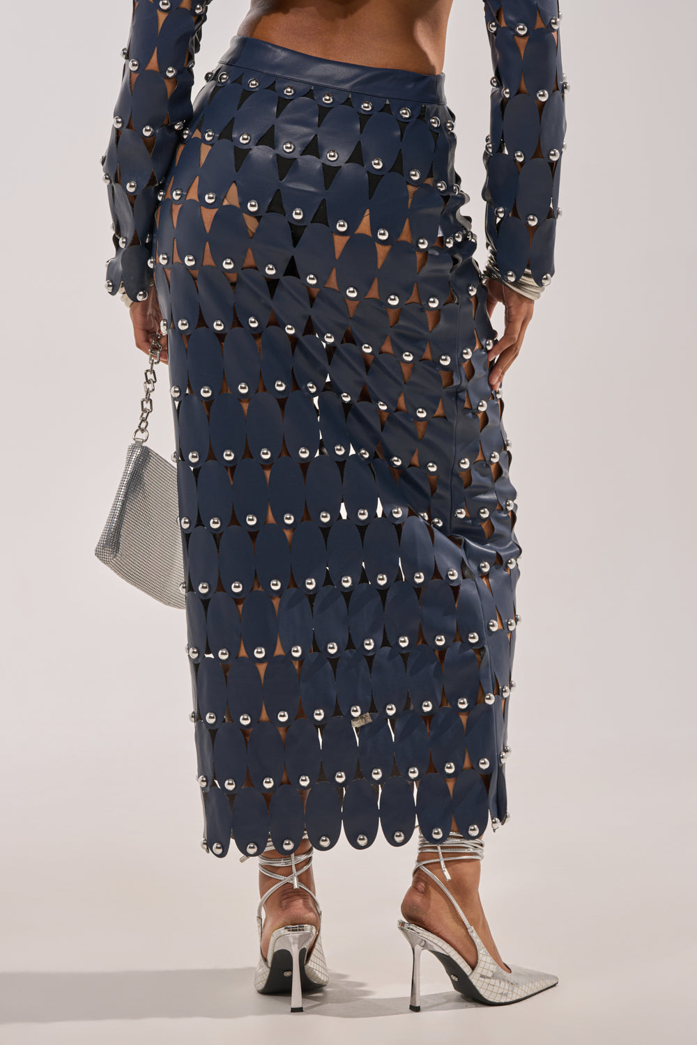 A person is seen from behind wearing THE SNEAK PEEK STUDDED PU MIDI SKIRT IN NAVY, featuring silver grommet accents and sheer cutouts, styled with a silver purse and silver high heels. Legs are visible through the skirt's fabric. 4