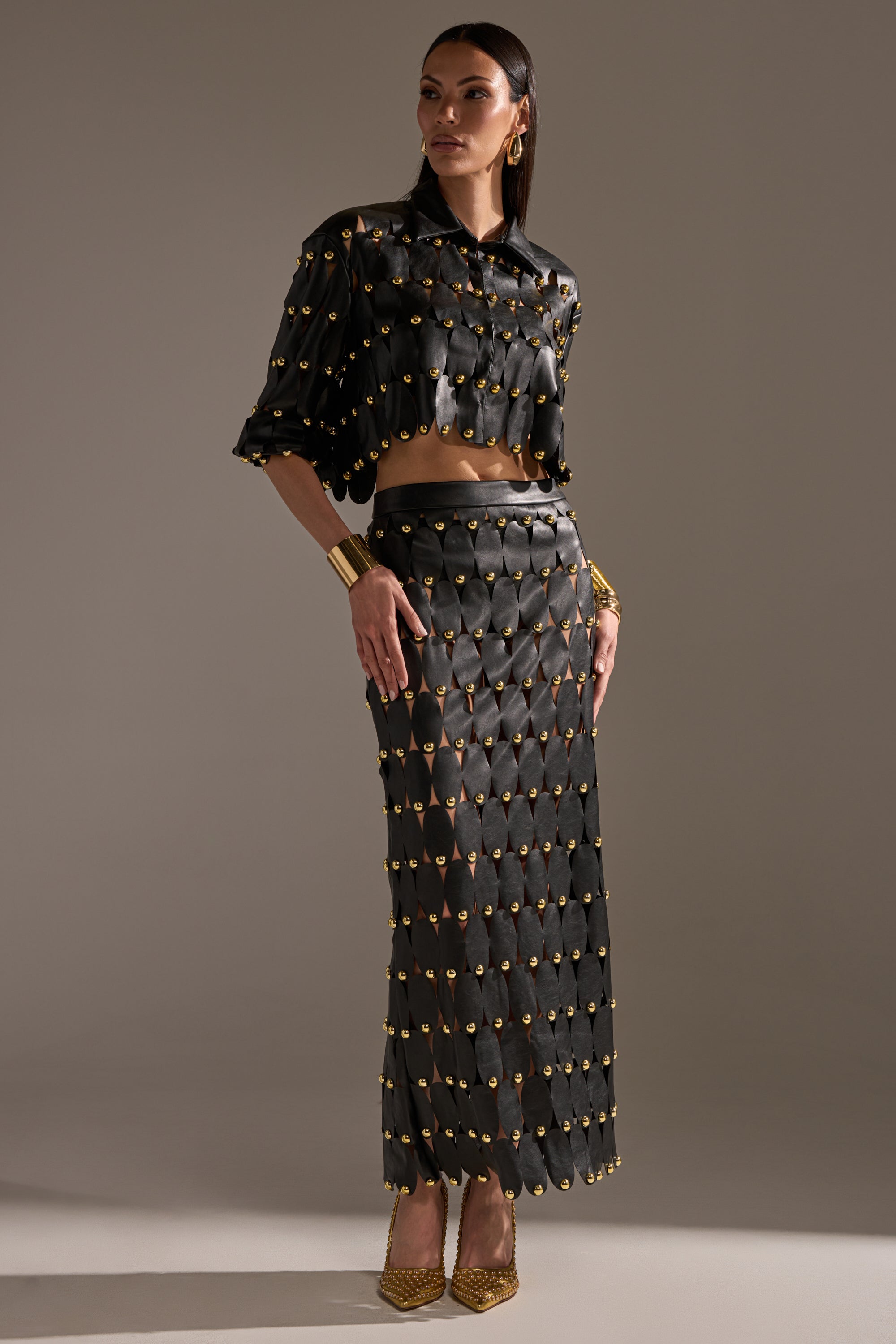 A woman poses in a high-fashion look featuring THE SNEAK PEEK STUDDED PU MIDI SKIRT IN BLACK, styled with a black cropped top, gold earrings and bracelets, and pointed heels against a plain background.