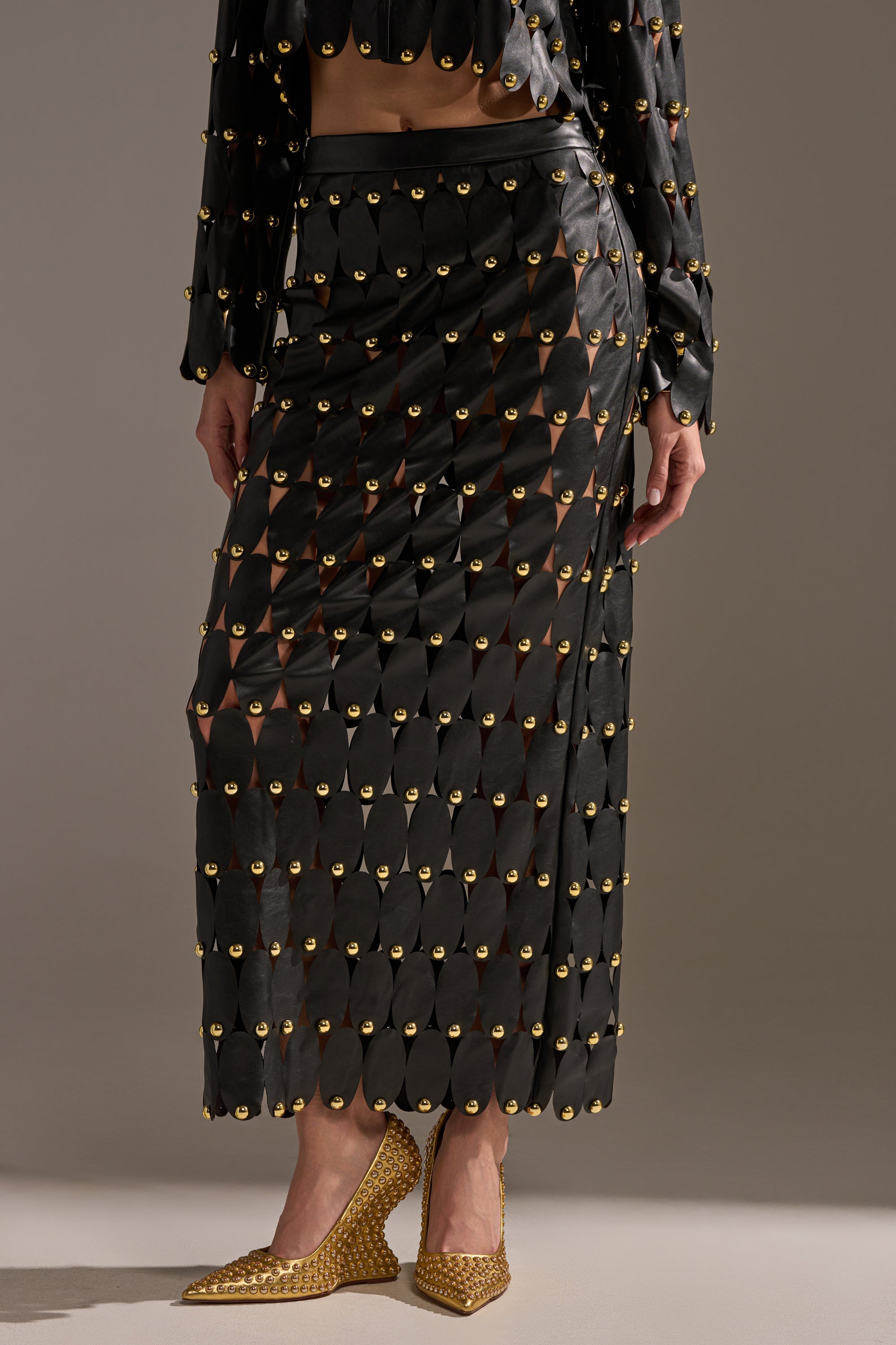 Wearing THE SNEAK PEEK STUDDED PU MIDI SKIRT IN BLACK with gold circular studs over a matching top and gold textured heels, the individual stands against a neutral background. The head is not visible.