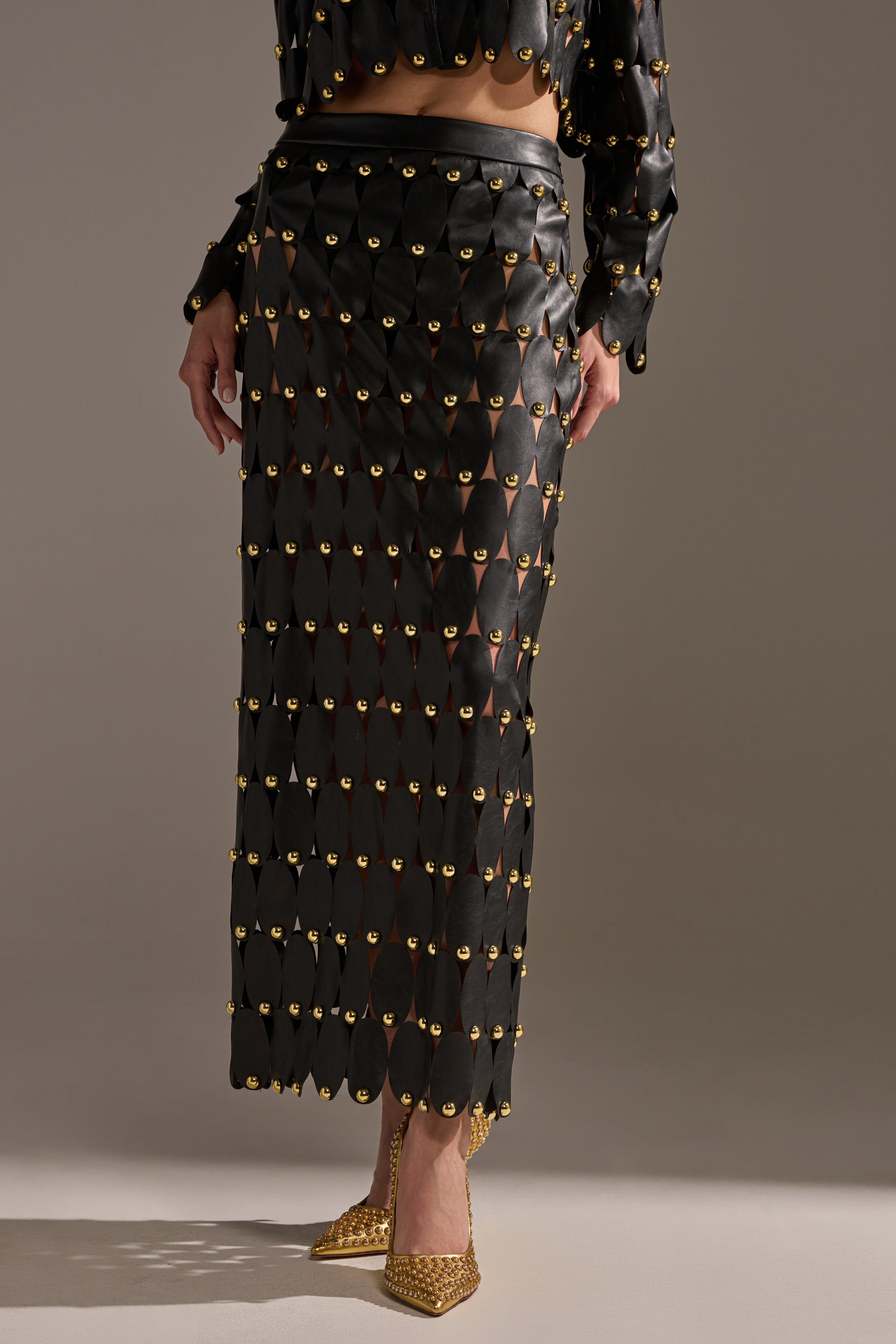 A person models THE SNEAK PEEK STUDDED PU MIDI SKIRT IN BLACK, featuring gold grommets and a geometric cut-out design with shiny gold accents, paired with matching pointed gold shoes. The upper body is not shown.