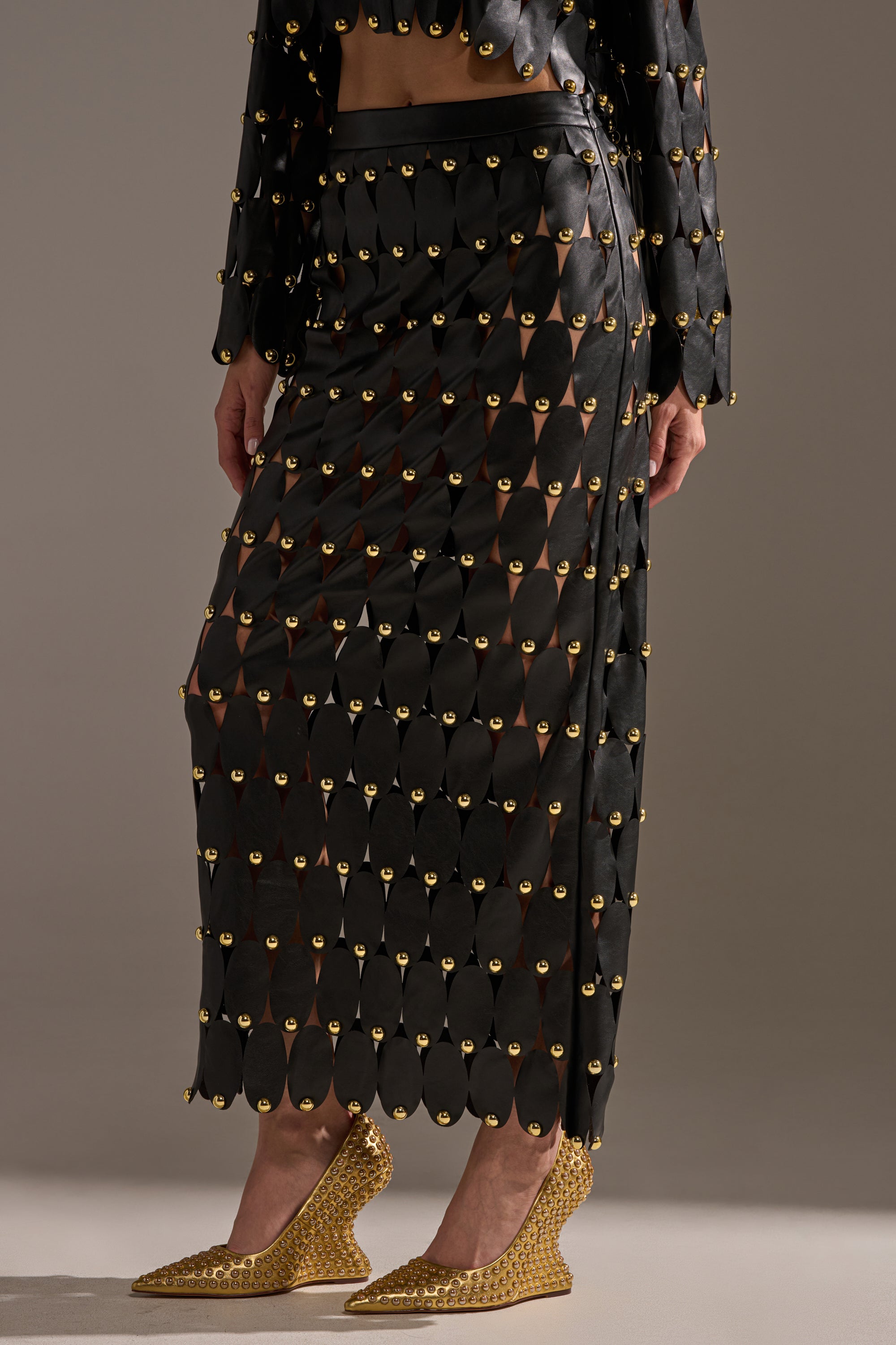 A person wears THE SNEAK PEEK STUDDED PU MIDI SKIRT IN BLACK, featuring gold embellishments and sheer panels, paired with textured gold pointed-toe shoes against a neutral background.