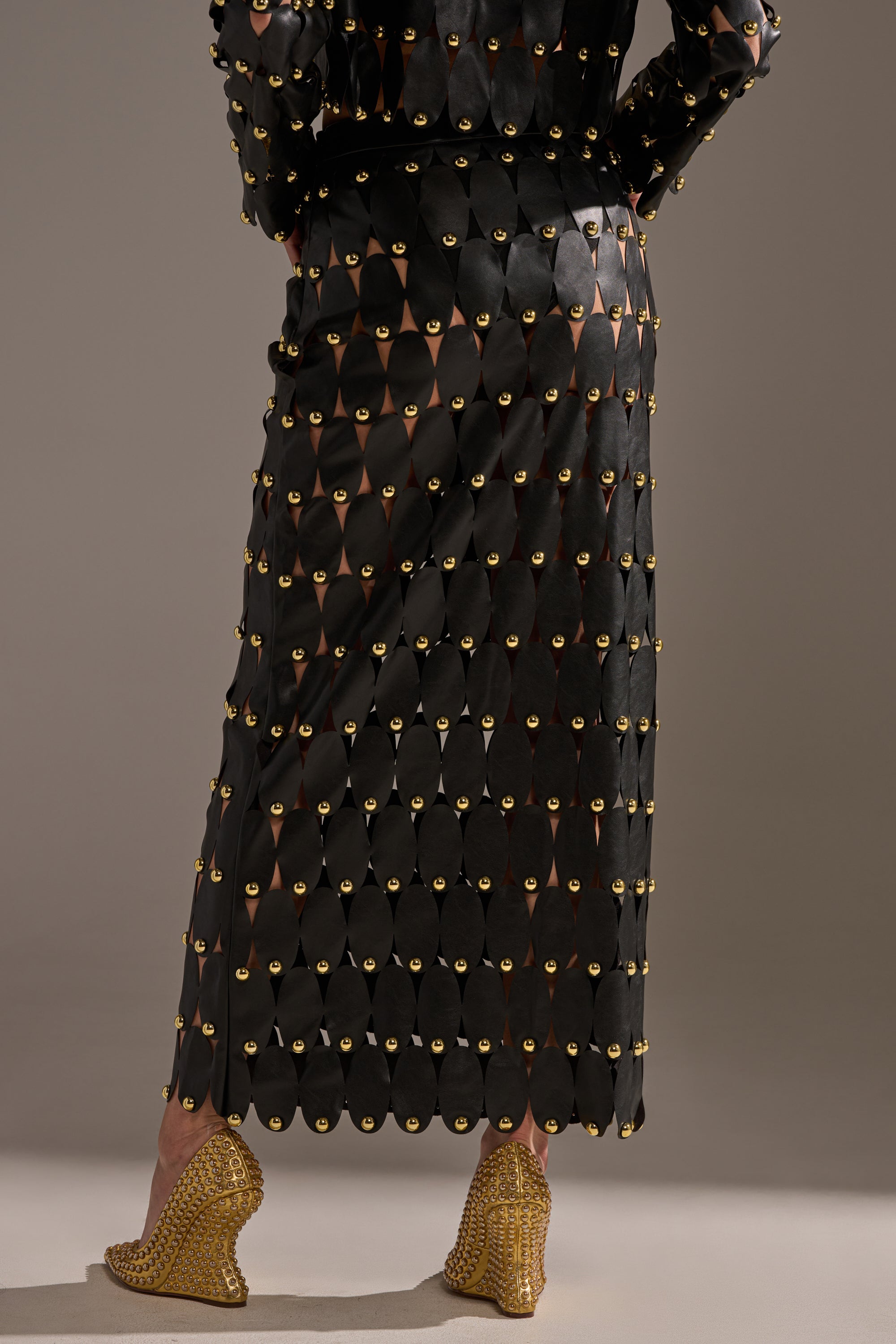 A person faces away, wearing THE SNEAK PEEK STUDDED PU MIDI SKIRT IN BLACK with scalloped edges and gold studs, styled with gold wedge heels against a plain, neutral background.
