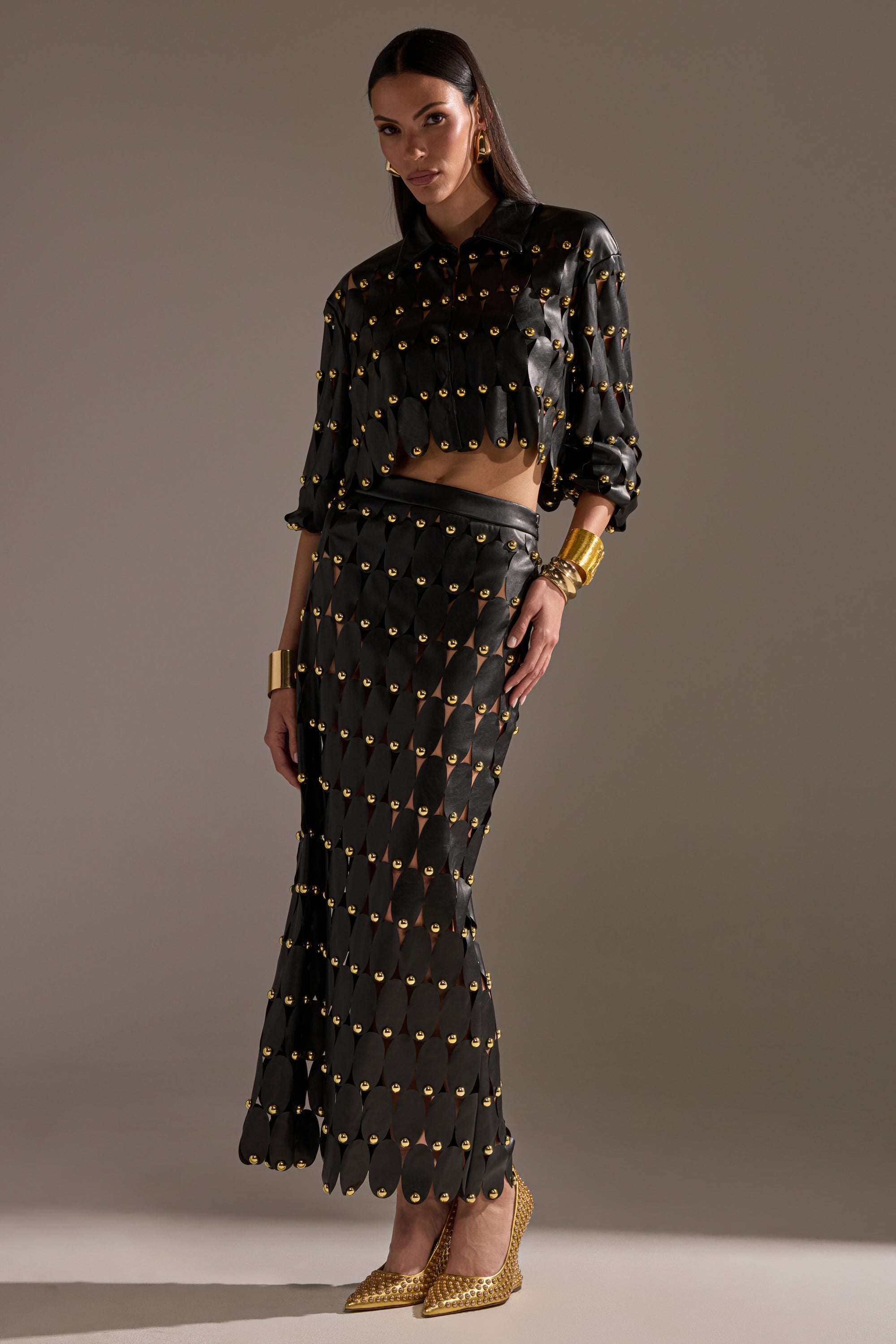 A woman poses in a black long-sleeved crop top and THE SNEAK PEEK STUDDED PU MIDI SKIRT IN BLACK, both with gold circular accents. She accessorizes with gold bracelets, pointed heels, and has sleek hair against a neutral background.