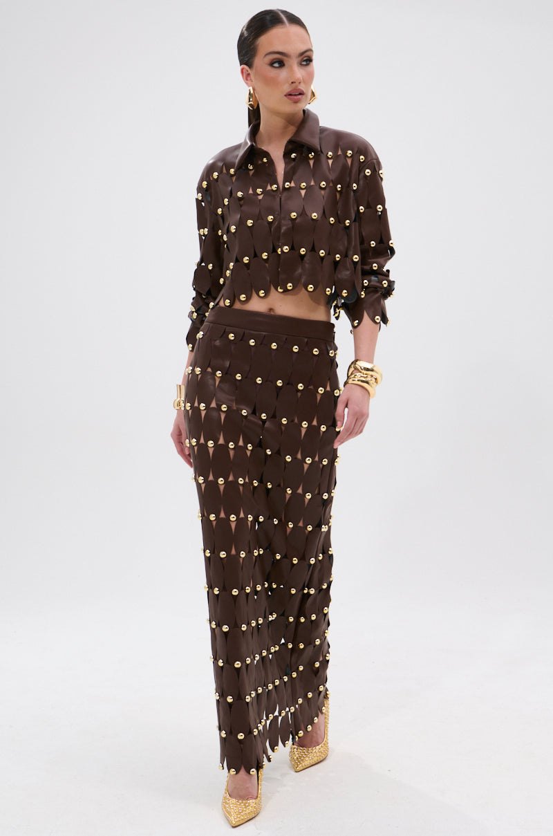 A woman models the SNEAK PEEK STUDDED PU MIDI SKIRT IN BROWN, featuring a high-rise waistband and gold circular studs, styled with gold accessories and beige heels against a plain white background.