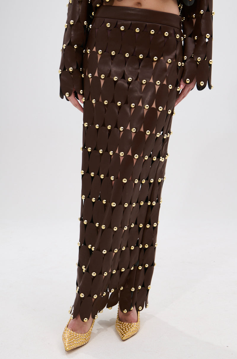 A person wears the SNEAK PEEK STUDDED PU MIDI SKIRT IN BROWN, featuring a high-rise waistband and overlapping oval panels, paired with gold pointed-toe shoes. The midriff is visible against a plain white background.