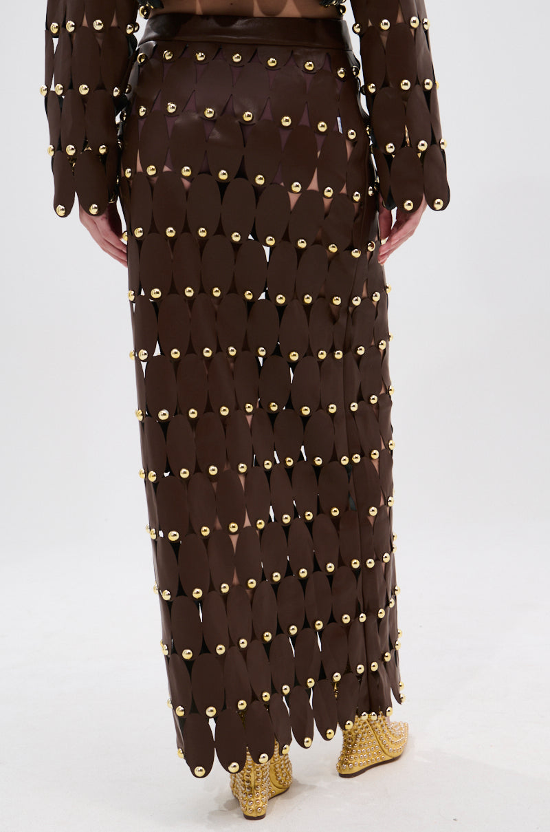 A person is shown from the back wearing the SNEAK PEEK STUDDED PU MIDI SKIRT IN BROWN, featuring overlapping oval pieces connected by gold rings, paired with gold embellished shoes against a plain white background.