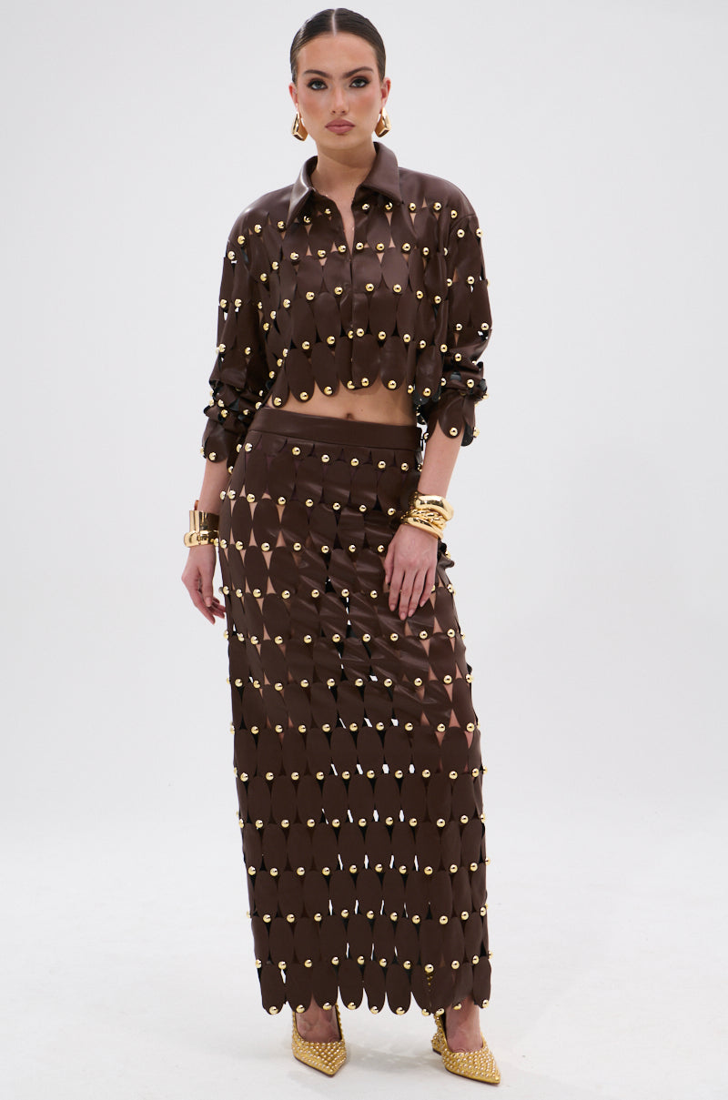A woman models the SNEAK PEEK STUDDED PU MIDI SKIRT IN BROWN, paired with a matching studded long-sleeve cropped top, gold jewelry, and gold heels. The skirt features circular studs and a high-rise waistband.