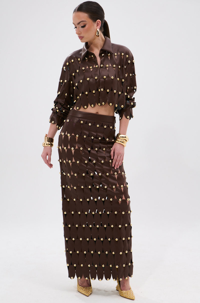 A woman wears the SNEAK PEEK STUDDED PU MIDI SKIRT IN BROWN with a matching long-sleeved crop top, gold earrings, bracelets, and pointed heels. The outfit features gold circular studs and a high-rise waistband against a plain background.