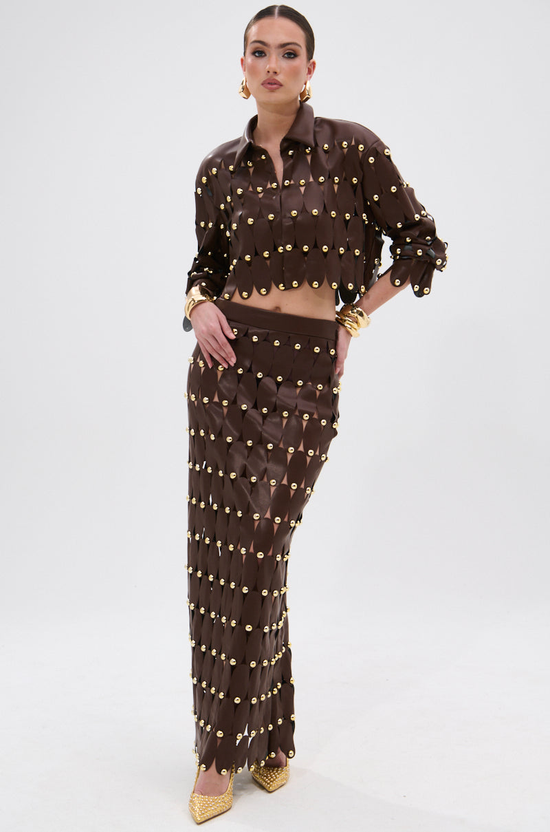 A woman models the SNEAK PEEK STUDDED PU MIDI SKIRT IN BROWN, paired with a matching cropped top. The look is finished with statement earrings, gold accessories, and pointed gold heels.