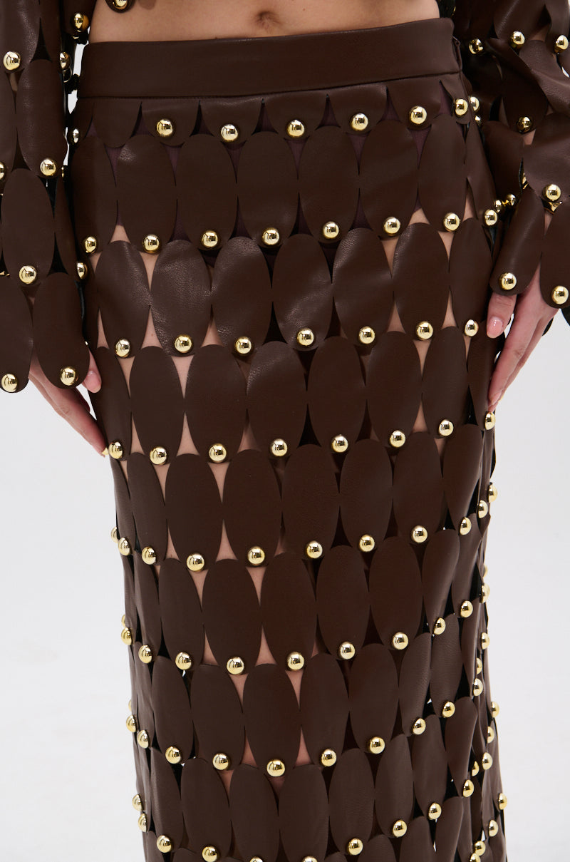 A close-up of the SNEAK PEEK STUDDED PU MIDI SKIRT IN BROWN, featuring overlapping oval panels with gold studs creating a textured grid pattern. The high-rise waistband and the wearer’s hands are partially visible at their sides.