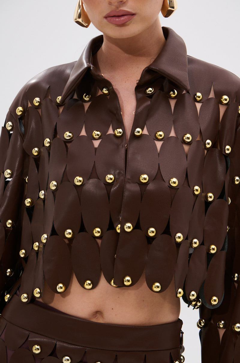 Wearing the SNEAK PEEK STUDDED PU CROP CARDIGAN IN BROWN, styled with matching bottoms and bold gold earrings for a modern, edgy vibe. The oval-shaped cutouts and metallic studs add unique detail, while the person's face is partially visible.