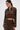 A woman in the SNEAK PEEK STUDDED PU CROP CARDIGAN IN BROWN and matching skirt, featuring metallic studs, stands against a plain background with her hair in a sleek ponytail and large gold hoop earrings.