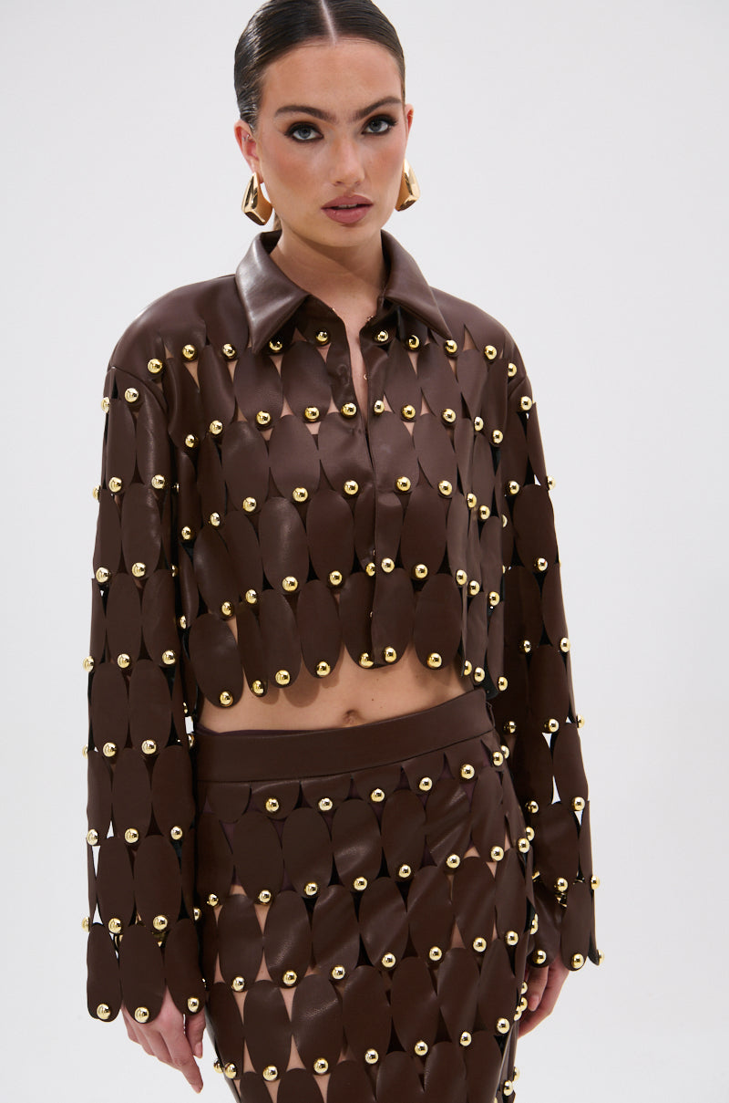 A woman models the SNEAK PEEK STUDDED PU CROP CARDIGAN IN BROWN, a faux leather piece with metallic studs, paired with a matching skirt. She stands against a white background, wearing slicked-back hair and gold hoop earrings.