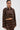 A woman models the SNEAK PEEK STUDDED PU CROP CARDIGAN IN BROWN, a faux leather piece with metallic studs, paired with a matching skirt. She stands against a white background, wearing slicked-back hair and gold hoop earrings.