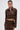 A woman wearing the SNEAK PEEK STUDDED PU CROP CARDIGAN IN BROWN with metallic stud details and a matching skirt poses against a white background, styled with slicked-back hair, bold makeup, and gold hoop earrings.