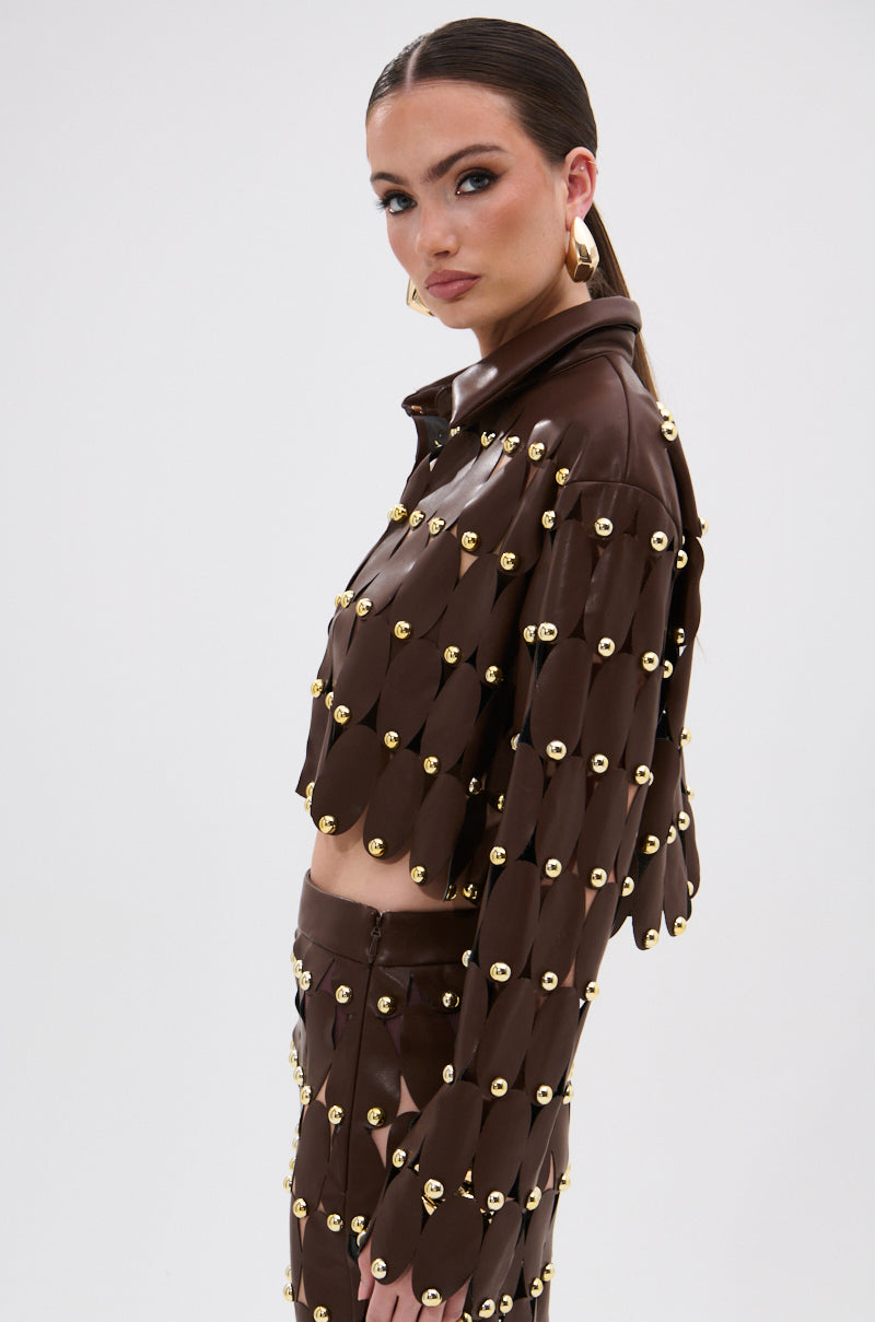 A woman wears the SNEAK PEEK STUDDED PU CROP CARDIGAN IN BROWN, styled with slicked-back hair, bold makeup, and large hoop earrings. She stands confidently against a white background, looking at the camera.
