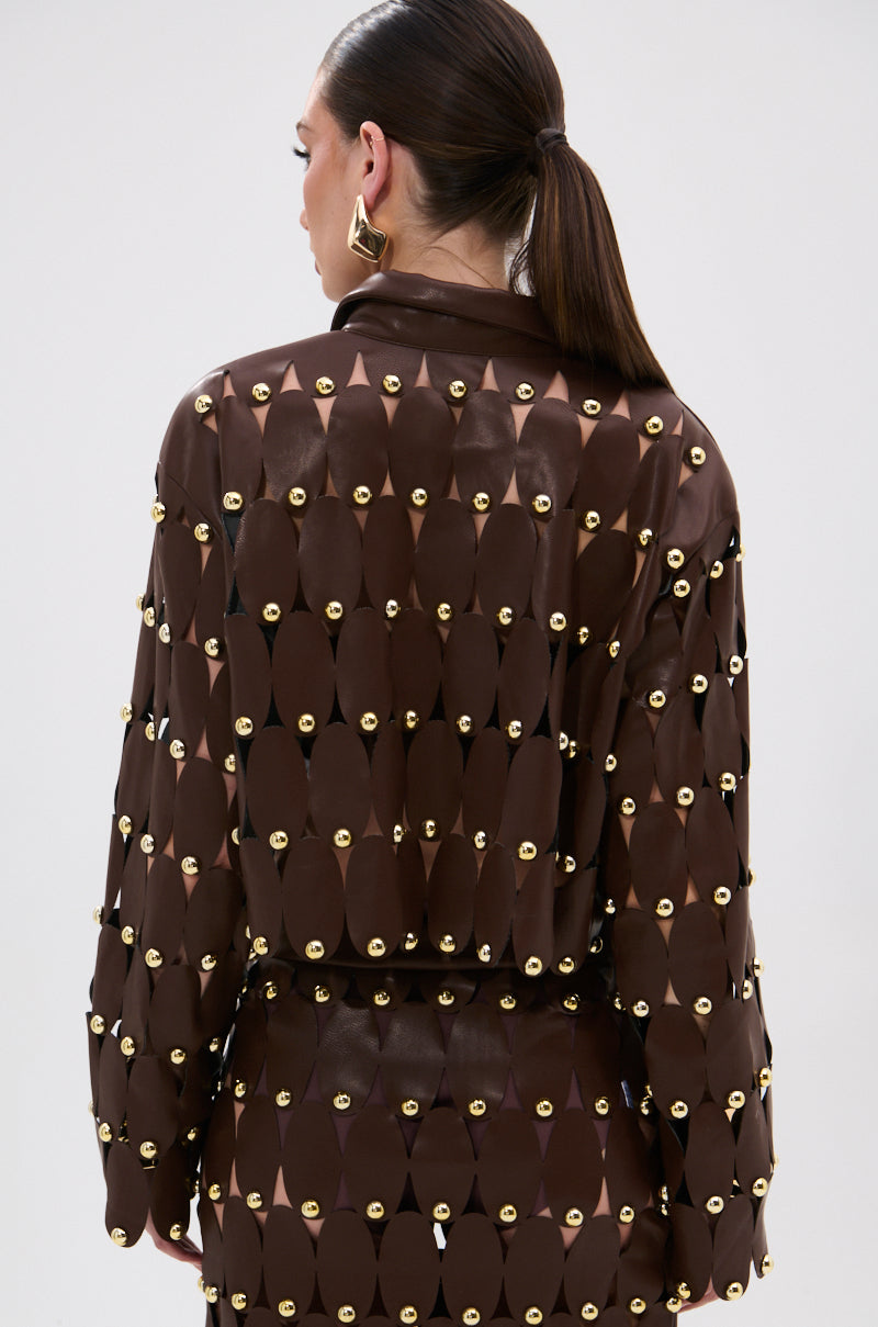 A woman with slicked-back hair is seen from the back wearing the SNEAK PEEK STUDDED PU CROP CARDIGAN IN BROWN, a sheer, long-sleeved top adorned with metallic studs, set against a plain light background.