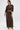 Wearing the SNEAK PEEK STUDDED PU CROP CARDIGAN IN BROWN and a matching studded long skirt, a woman stands against a plain white background, accessorized with gold earrings, bracelets, and heels, looking down and to the side.