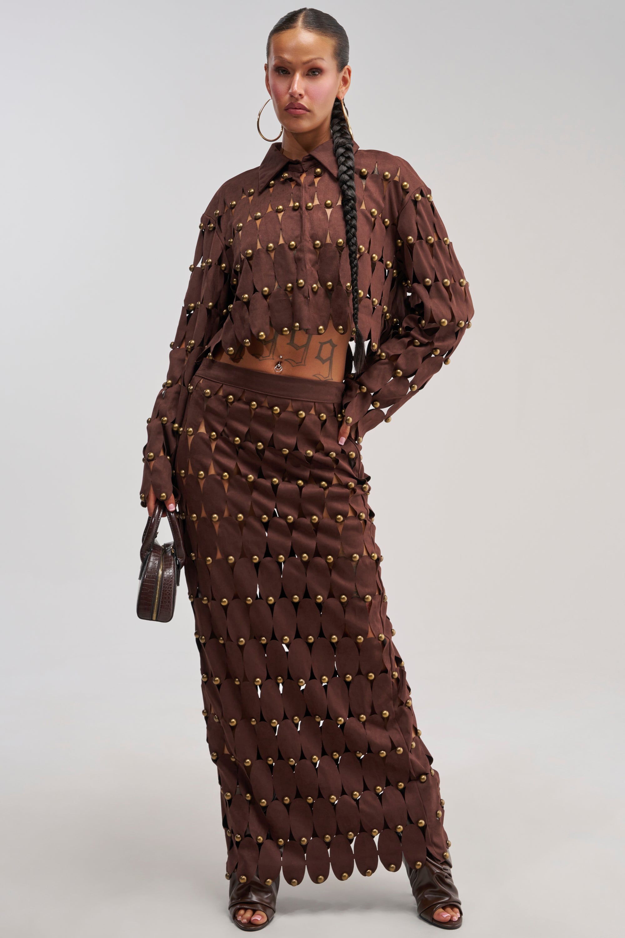 A woman poses in THE SNEAK PEEK STUDDED SUEDE MIDI SKIRT, a brown midi skirt with gold embellishments, styled with a long-sleeve button-up top. She has a long braid, hoop earrings, carries a small brown handbag, and wears open-toe heels.