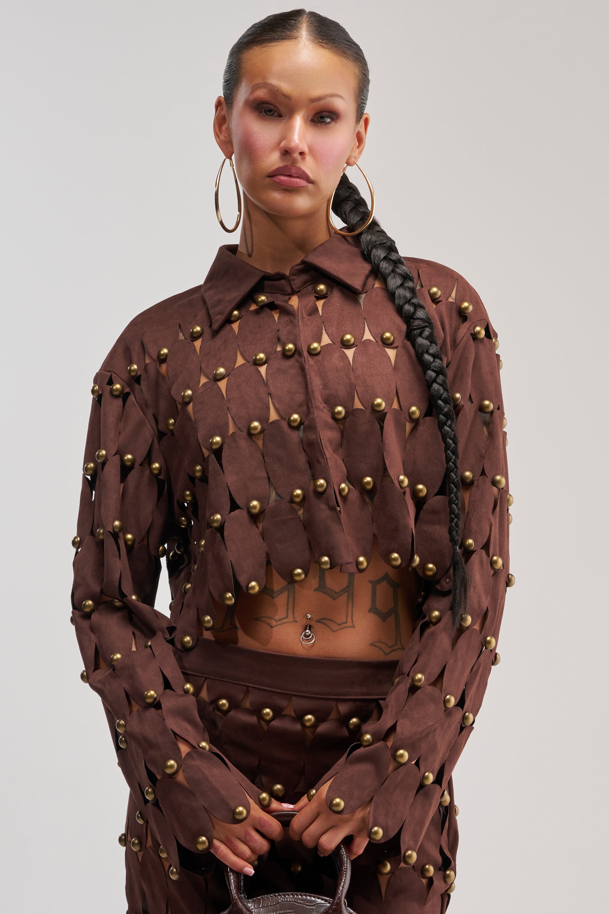 A woman with a long braid models THE SNEAK PEEK STUDDED SUEDE BLOUSE, a brown suede top embellished with gold studs, styled with large hoop earrings and showcasing tattoos on her torso, standing against a plain background.