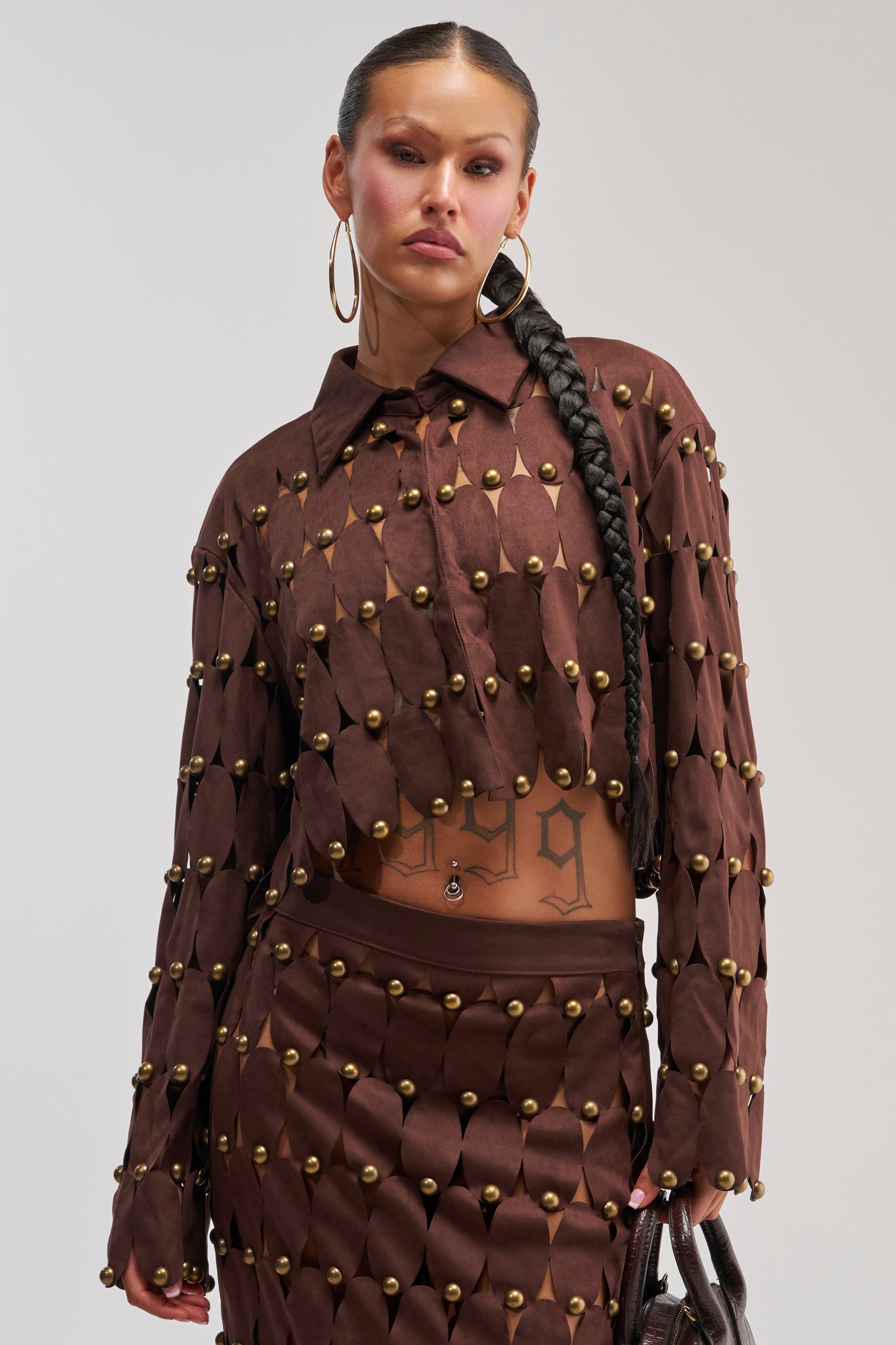 A person with a long braided ponytail wears THE SNEAK PEEK STUDDED SUEDE BLOUSE as part of a matching textured two-piece outfit accented with gold studs. They accessorize with large hoop earrings and carry a bag, revealing a tattoo on their midriff.