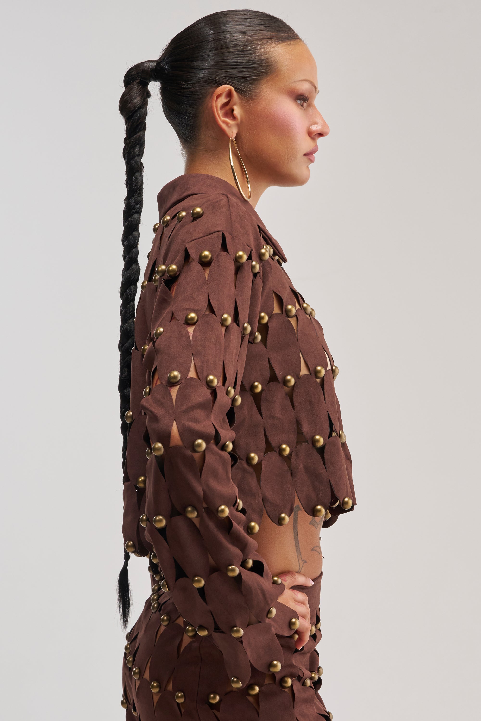 Wearing THE SNEAK PEEK STUDDED SUEDE BLOUSE, a woman with a long braided ponytail and large hoop earrings stands in profile against a plain light background; the brown suede blouse is adorned with gold studs.