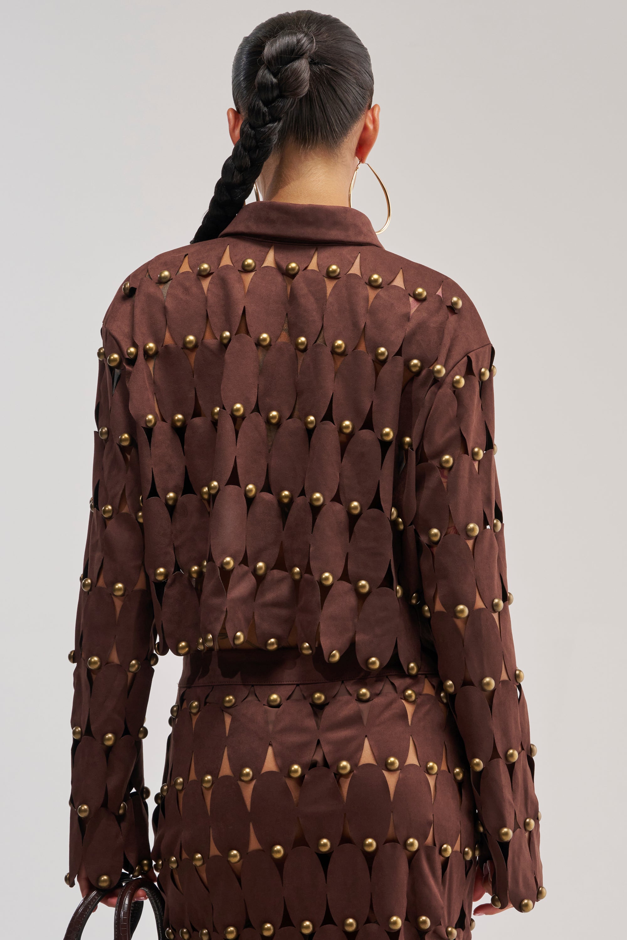 A person with a long braided ponytail is seen from behind wearing THE SNEAK PEEK STUDDED SUEDE BLOUSE paired with a matching skirt, both featuring oval cutouts and gold studs. They accessorize with large hoop earrings and hold a small bag.