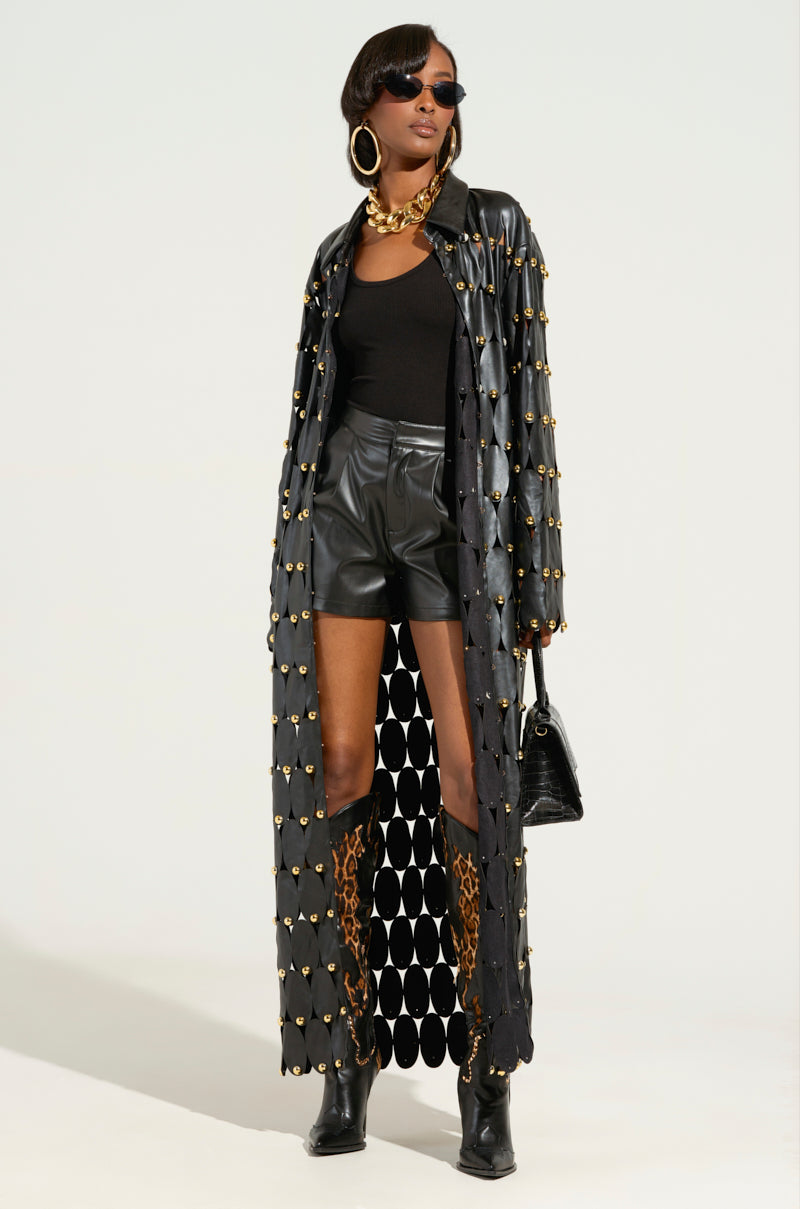 A person showcases "THE SNEAK PEEK STUDDED PU LONG CARDIGAN IN BLACK" adorned with metallic studs, along with black shorts, a sleek top, and knee-high leopard print boots. They complement the outfit with sunglasses, large hoop earrings, and a gold necklace while holding an elegant black handbag. 0