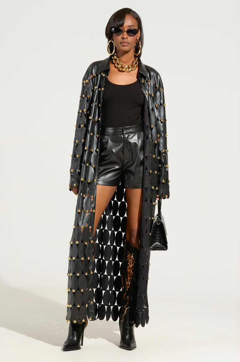 A person is dressed in THE SNEAK PEEK STUDDED PU LONG CARDIGAN IN BLACK, layered over a black top and leather shorts. They accessorize with oversized sunglasses, gold hoop earrings, and a chunky necklace. Completing the look, they carry a small black handbag and wear knee-high boots featuring leopard print accents. 1