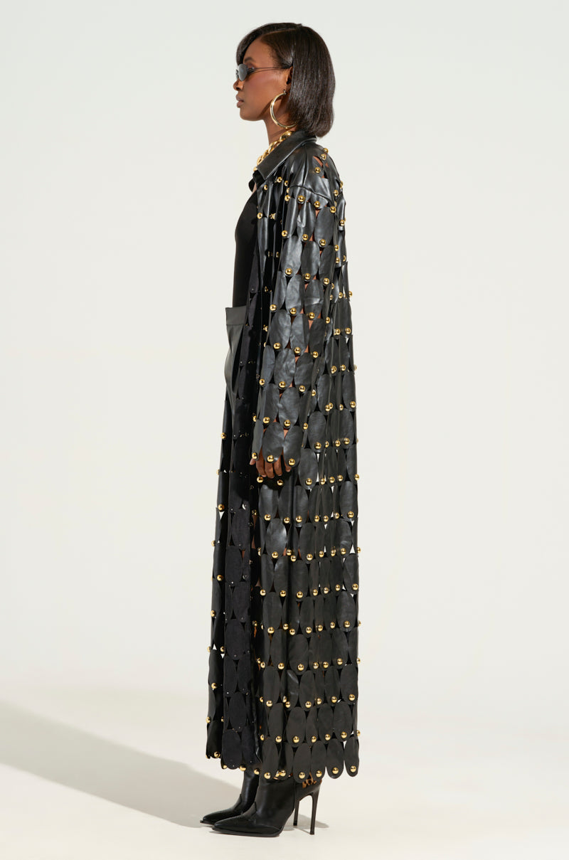 A person dressed in THE SNEAK PEEK STUDDED PU LONG CARDIGAN IN BLACK, featuring metallic stud embellishments and a scalloped design, stands in profile view with high-heeled boots. They accessorize their look with sunglasses and large hoop earrings, complementing their short haircut. 2
