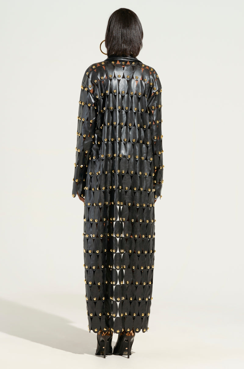 A person in THE SNEAK PEEK STUDDED PU LONG CARDIGAN IN BLACK, distinguished by its metallic stud embellishments and a unique circular pattern, stands with their back to the camera, complemented by high heels. 3