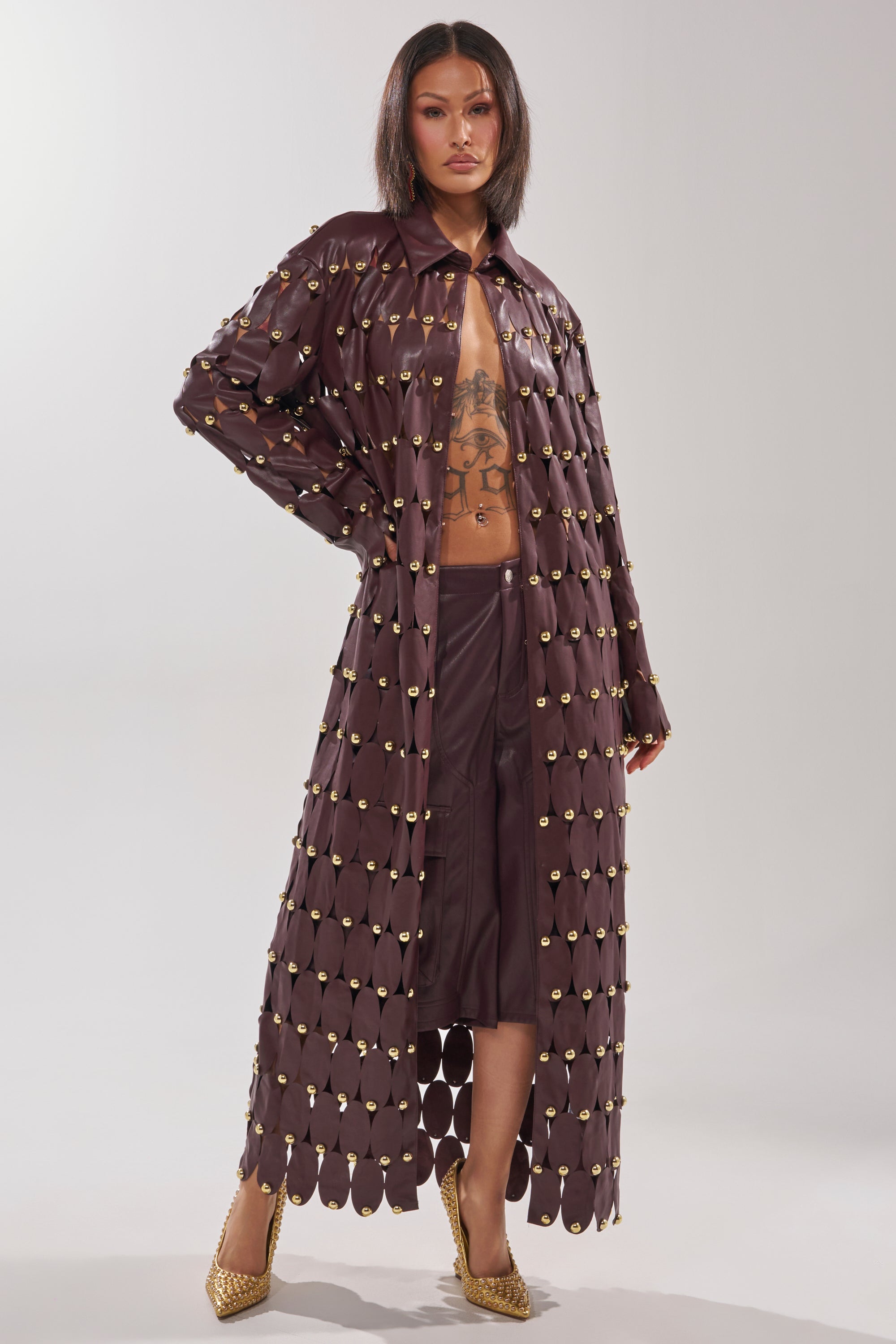 A woman stands confidently in THE SNEAK PEEK STUDDED PU LONG CARDIGAN IN BURGUNDY, styled open to reveal her tattooed torso and matching shorts. Gold circular accents and gold high heels complete her bold look.
