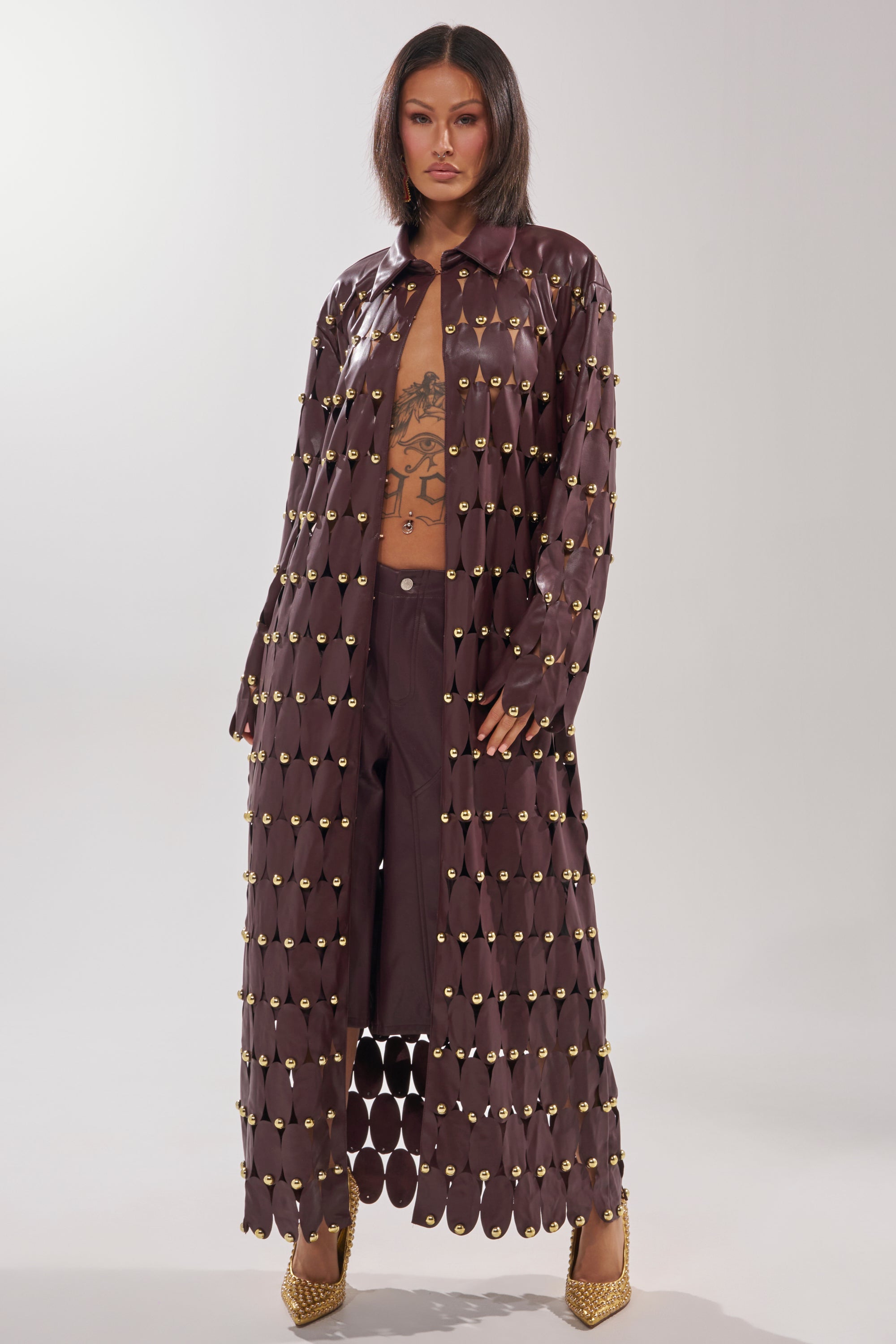 A woman stands confidently in THE SNEAK PEEK STUDDED PU LONG CARDIGAN IN BURGUNDY with gold accents over matching pants. She wears gold high heels, has straight dark hair, visible torso tattoos, and poses against a plain light background.