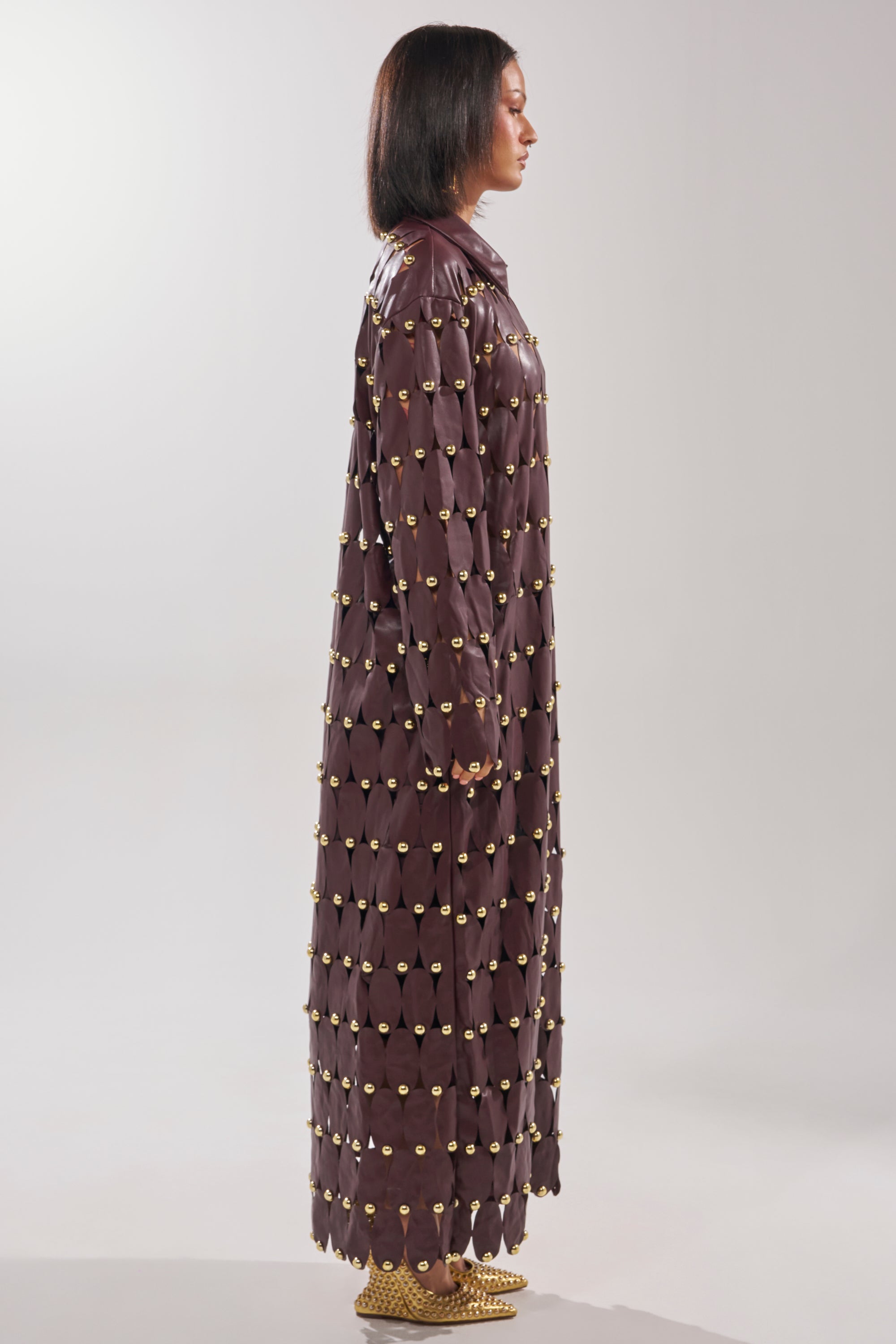 A person stands in profile against a light background wearing THE SNEAK PEEK STUDDED PU LONG CARDIGAN IN BURGUNDY, featuring rows of gold studs. The long, dark burgundy faux leather coat reaches the ankles and is styled with straight, shoulder-length hair.