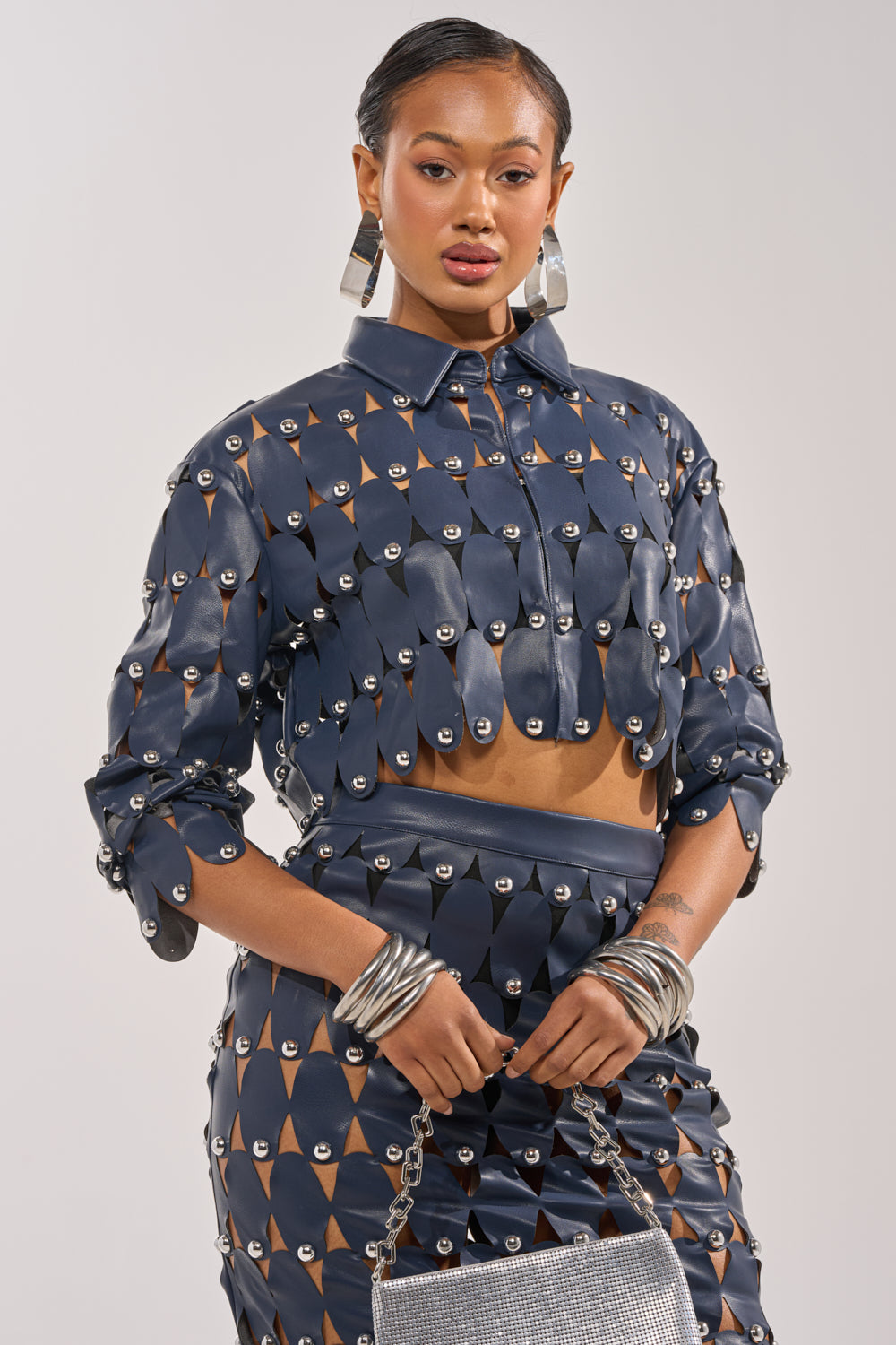 A woman models THE SNEAK PEEK STUDDED PU CROP CARDIGAN IN NAVY over a geometric-patterned crop top and skirt, accessorized with hoop earrings, chunky bangles, and a silver textured handbag against a neutral background. 1