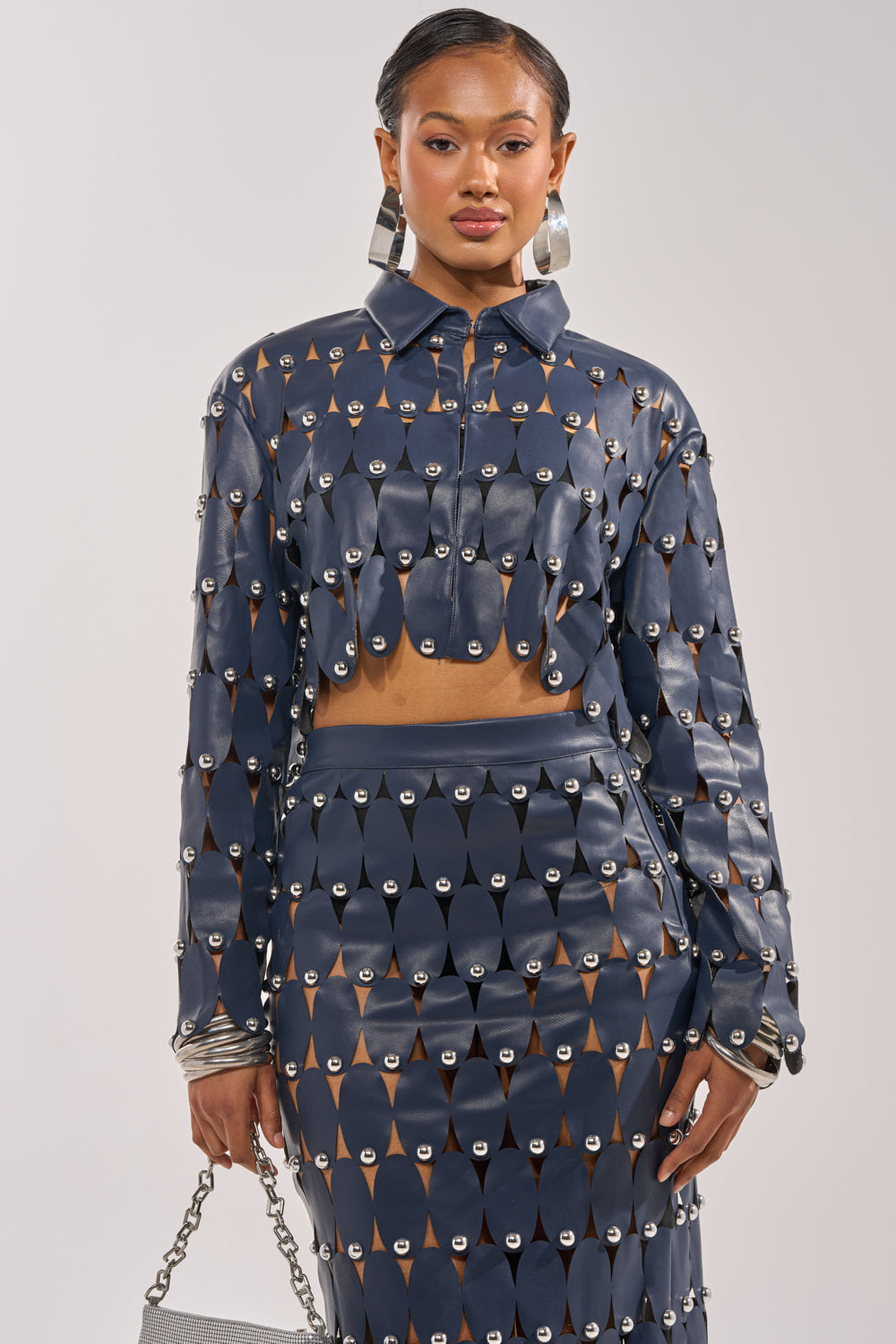 A woman wears THE SNEAK PEEK STUDDED PU CROP CARDIGAN IN NAVY over a navy crop top and skirt with silver grommets. She completes the look with a silver chain handbag and hoop earrings, posing against a light background. 2
