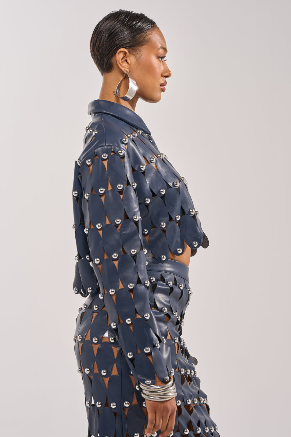 A woman in profile models THE SNEAK PEEK STUDDED PU CROP CARDIGAN IN NAVY, featuring geometric cut-outs and silver studs. She pairs it with large hoop earrings, silver bangles, and wears her short hair sleek against a plain backdrop. 3