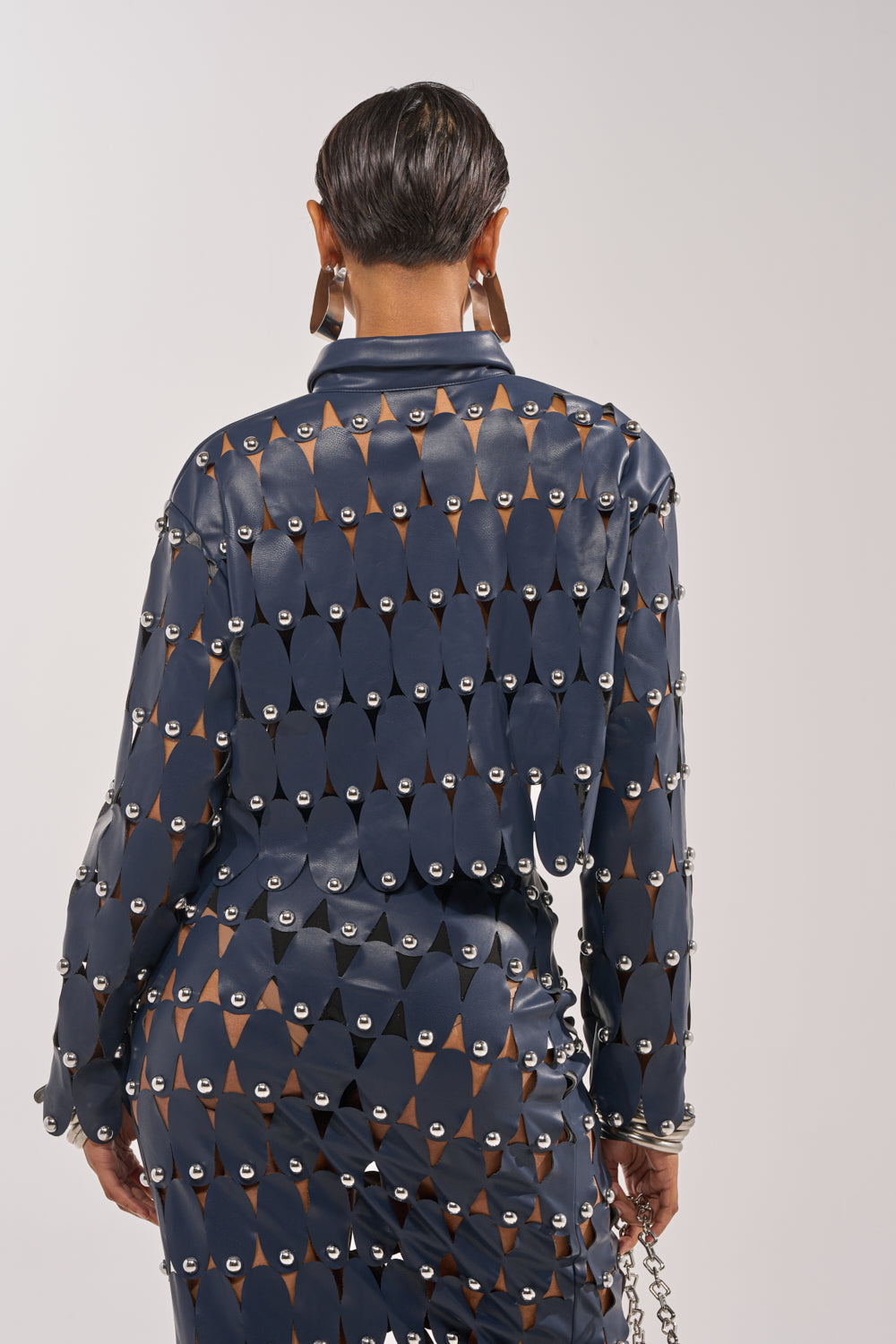 A person with short dark hair is seen from behind wearing THE SNEAK PEEK STUDDED PU CROP CARDIGAN IN NAVY, featuring a shiny finish and metallic details, holding a silver chain accessory against a plain light background. 4