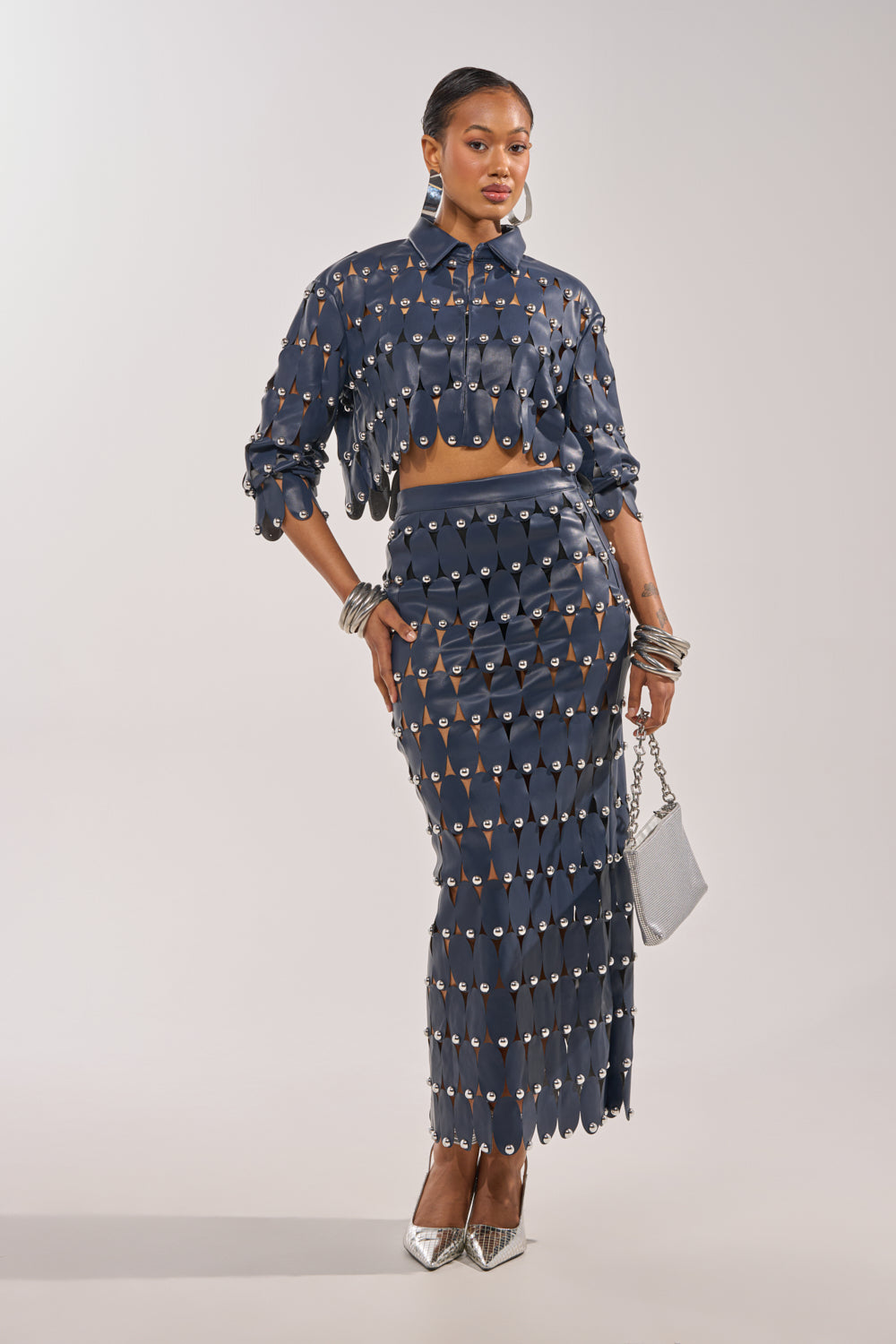 A woman stands confidently in a dark blue two-piece with cutouts, silver jewelry, metallic heels, and THE SNEAK PEEK STUDDED PU CROP CARDIGAN IN NAVY draped over her shoulders, carrying a small silver handbag. 6