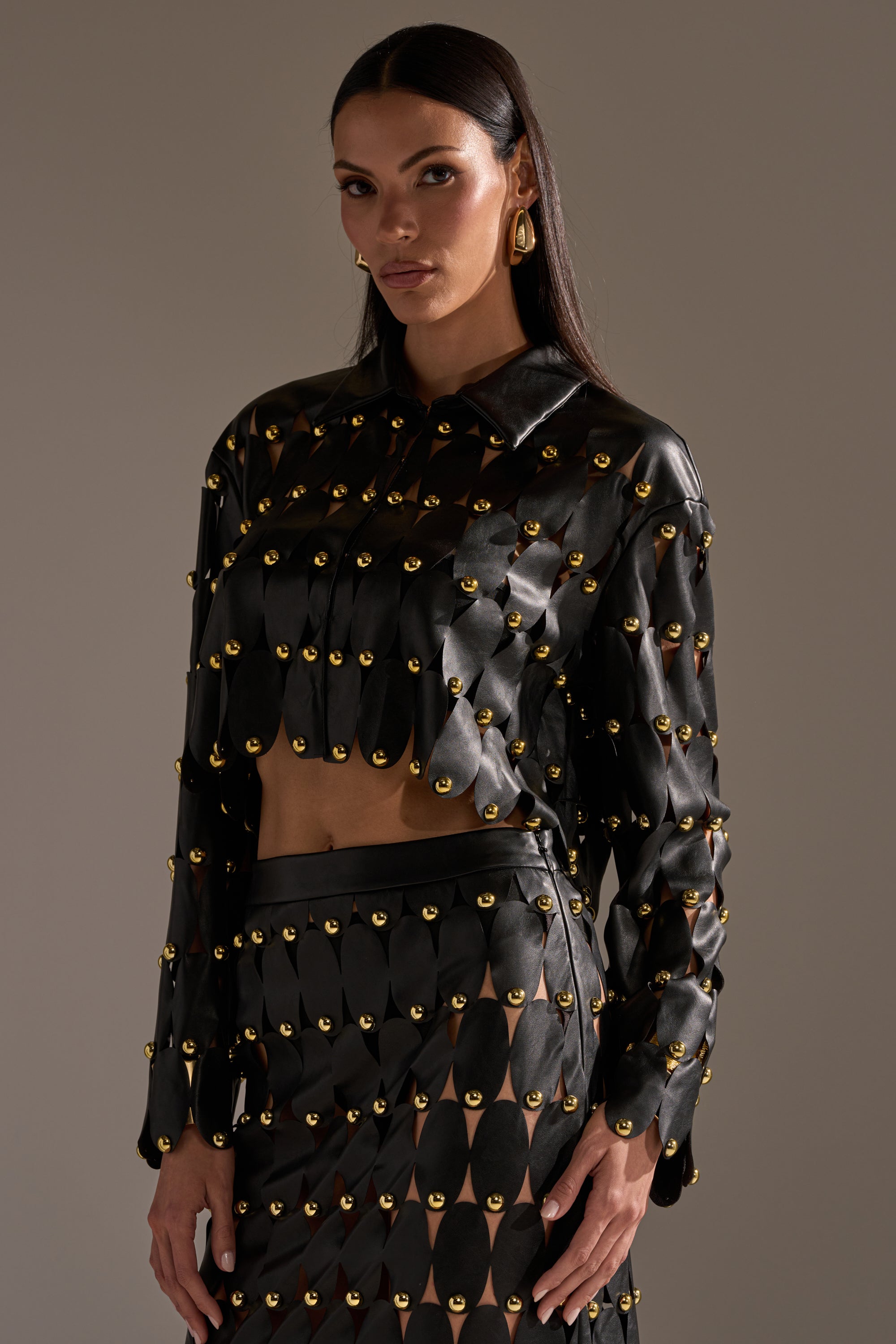 A woman wears THE SNEAK PEEK STUDDED PU BLOUSE IN BLACK, paired with a matching studded skirt. Her long, straight dark hair and gold earrings complete the look as she stands against a plain, neutral background.