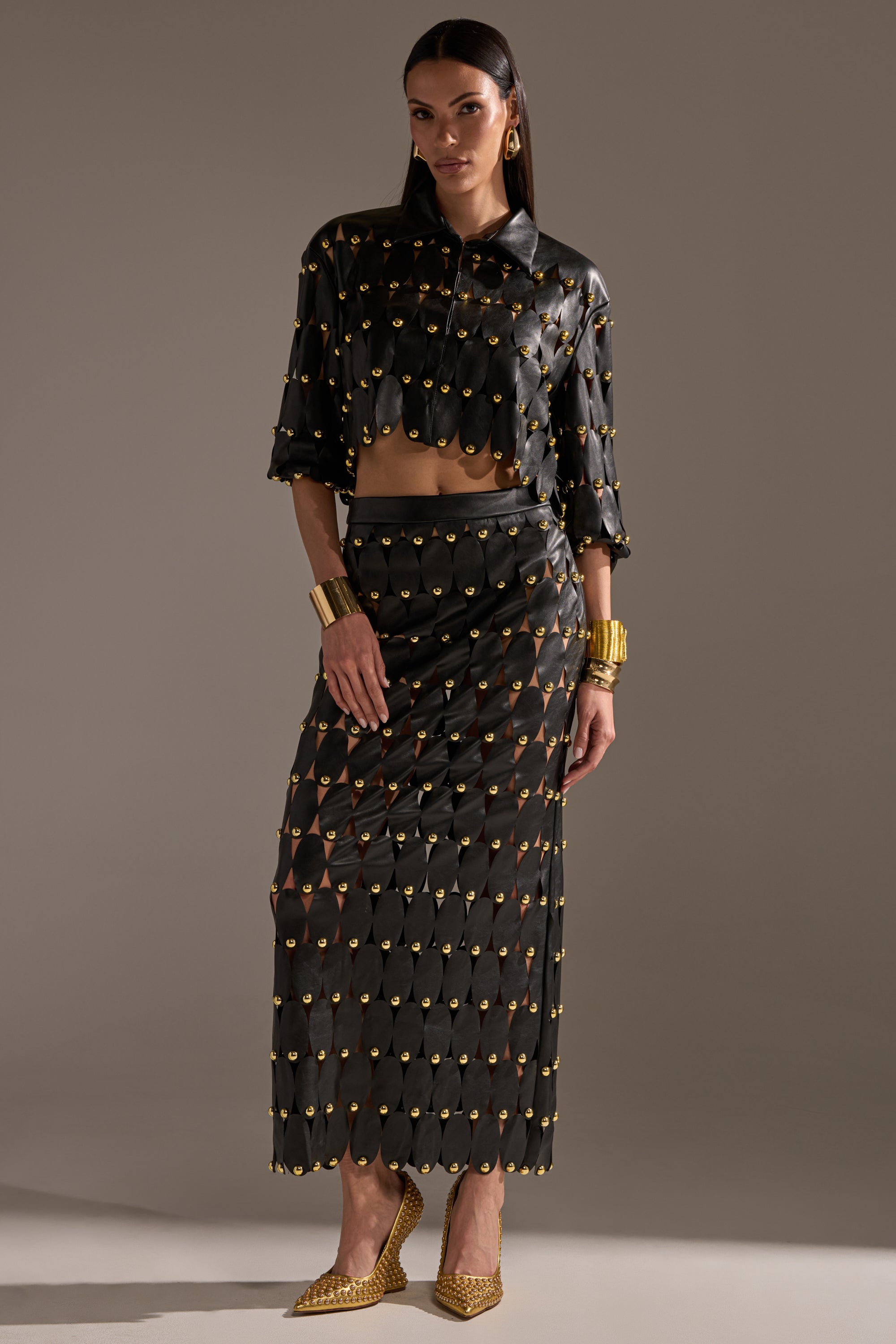 A woman wears THE SNEAK PEEK STUDDED PU BLOUSE IN BLACK, a sheer, cropped AKIRA Label top with gold embellishments, paired with an ankle-length skirt. She completes the look with gold bracelets and pointed gold heels. 9