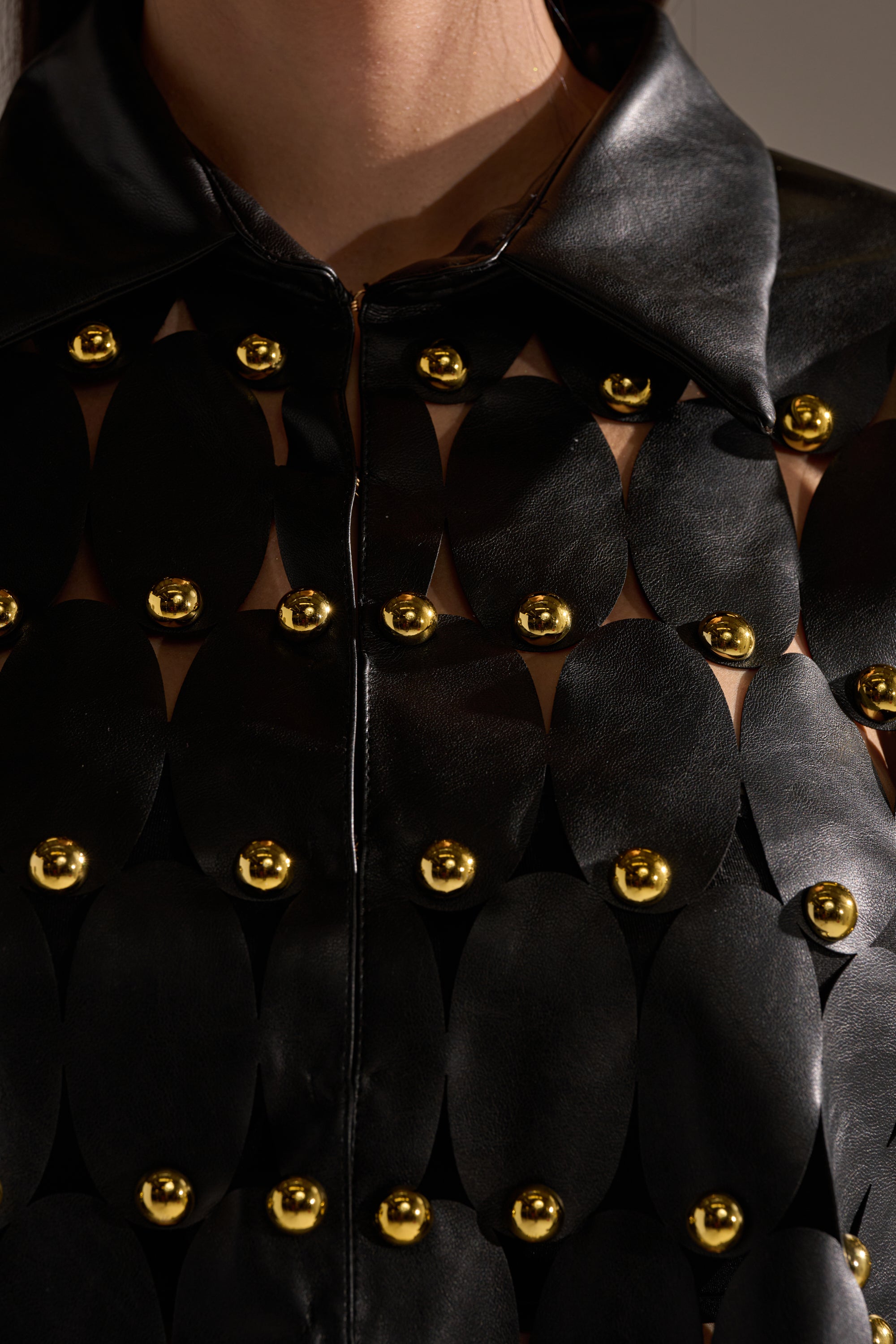 Close-up of THE SNEAK PEEK STUDDED PU BLOUSE IN BLACK, featuring oval cutouts linked by gold studs, creating a textured geometric pattern on the black PU fabric. 11