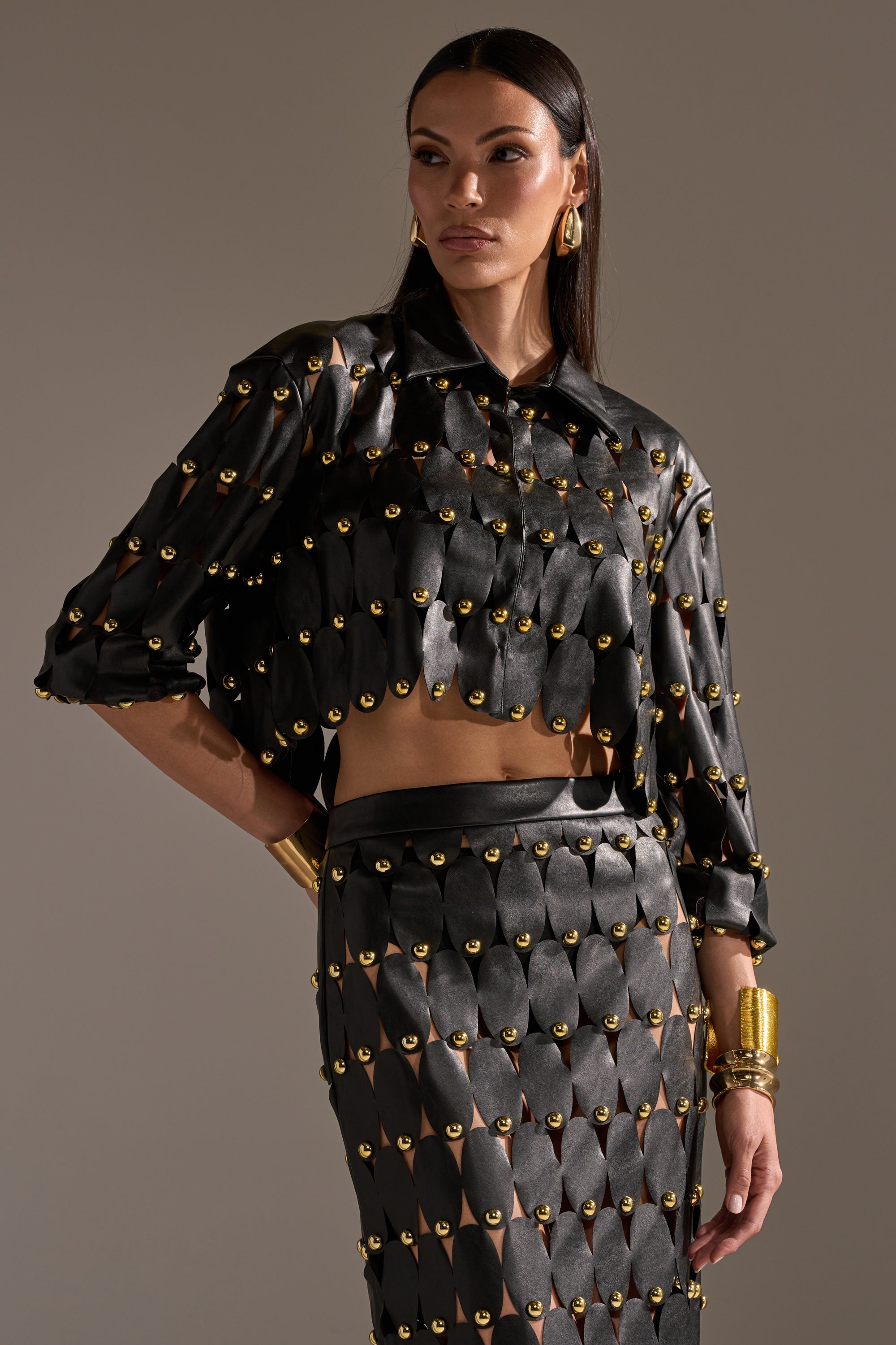 A woman wears THE SNEAK PEEK STUDDED PU BLOUSE IN BLACK by AKIRA Label, styled with matching black skirt, chunky gold earrings, and bracelets as she stands confidently against a neutral background. 1