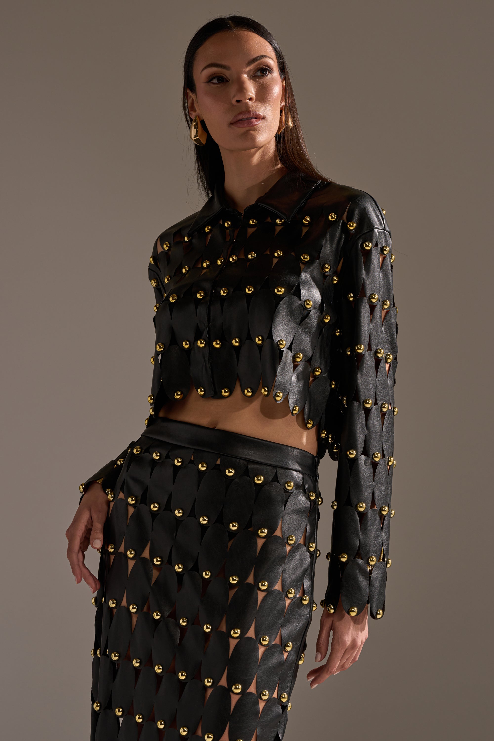 A woman poses confidently against a neutral background, wearing THE SNEAK PEEK STUDDED PU BLOUSE IN BLACK paired with a matching studded skirt. 2