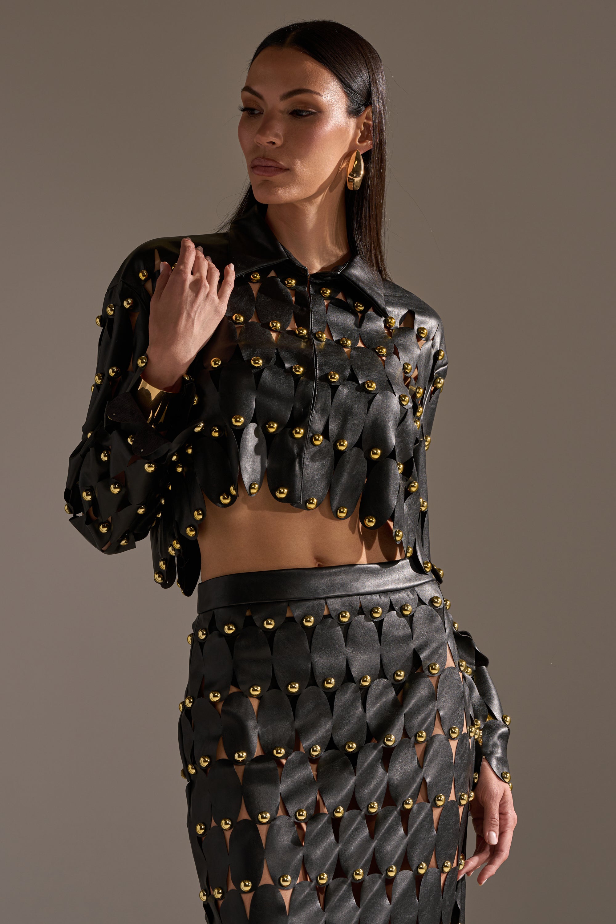 A woman models THE SNEAK PEEK STUDDED PU BLOUSE IN BLACK by AKIRA Label with long, straight hair and gold hoop earrings. The black blouse features studded details for a chic, modern look. 3