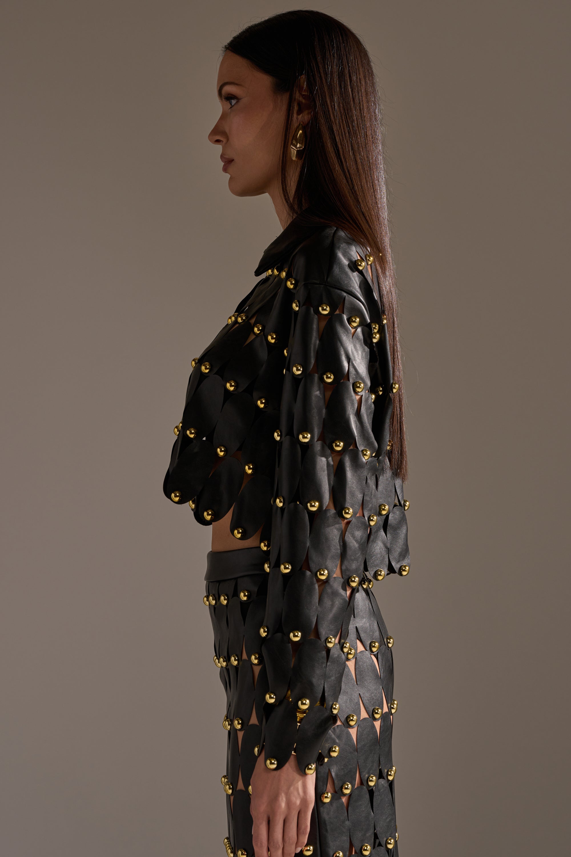 A woman in profile wears THE SNEAK PEEK STUDDED PU BLOUSE IN BLACK, featuring evenly spaced gold studs. Her straight, long hair and gold earrings accentuate the blouse’s bold metallic details against a simple backdrop. 4